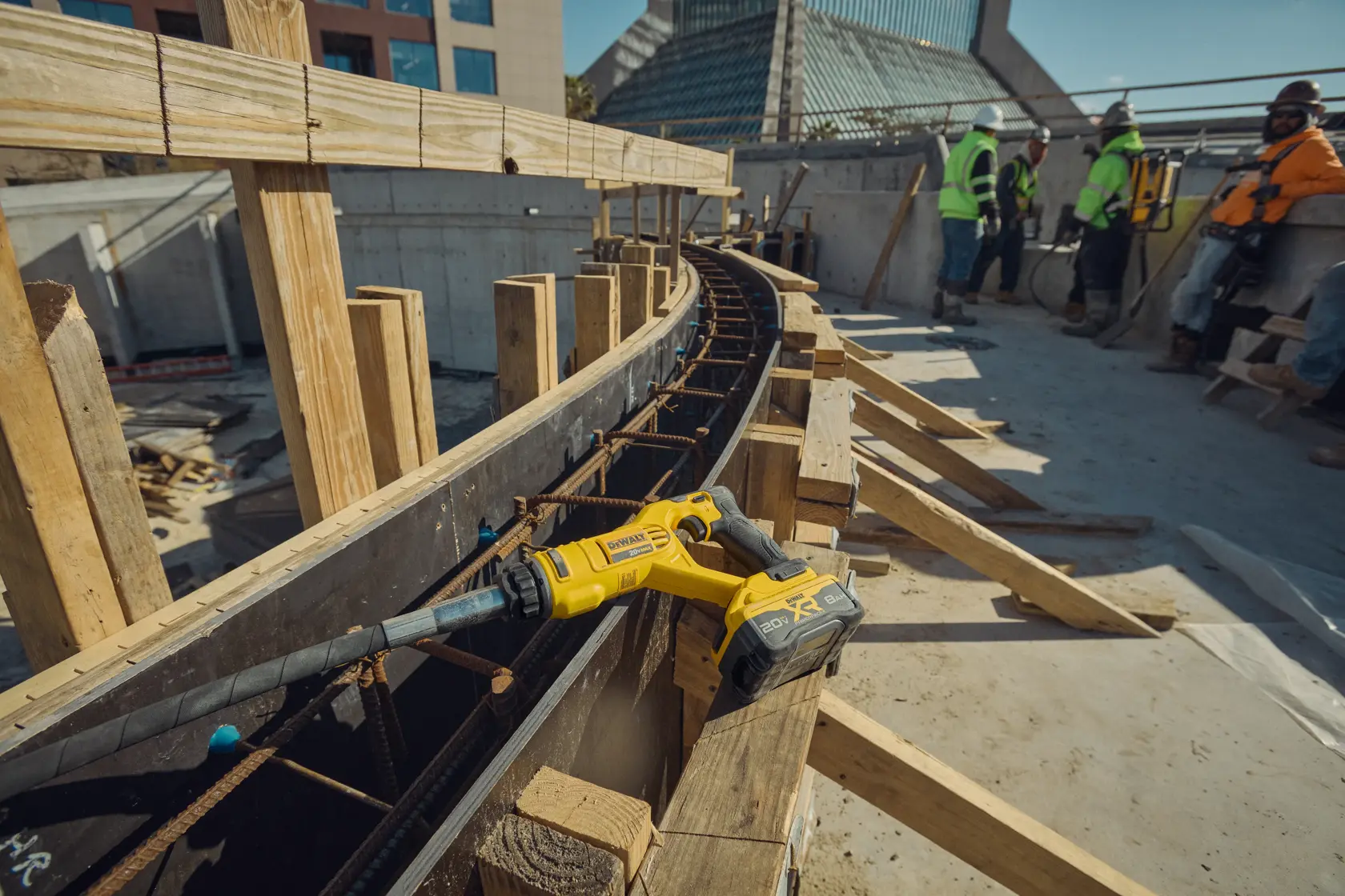 Two construction workers wearing safety helmets and orange high-visibility shirts are working at a construction site. One worker is using a DEWALT concrete vibrator tool on freshly poured concrete, while the other directs a concrete hose. Modern office buildings are visible in the background.