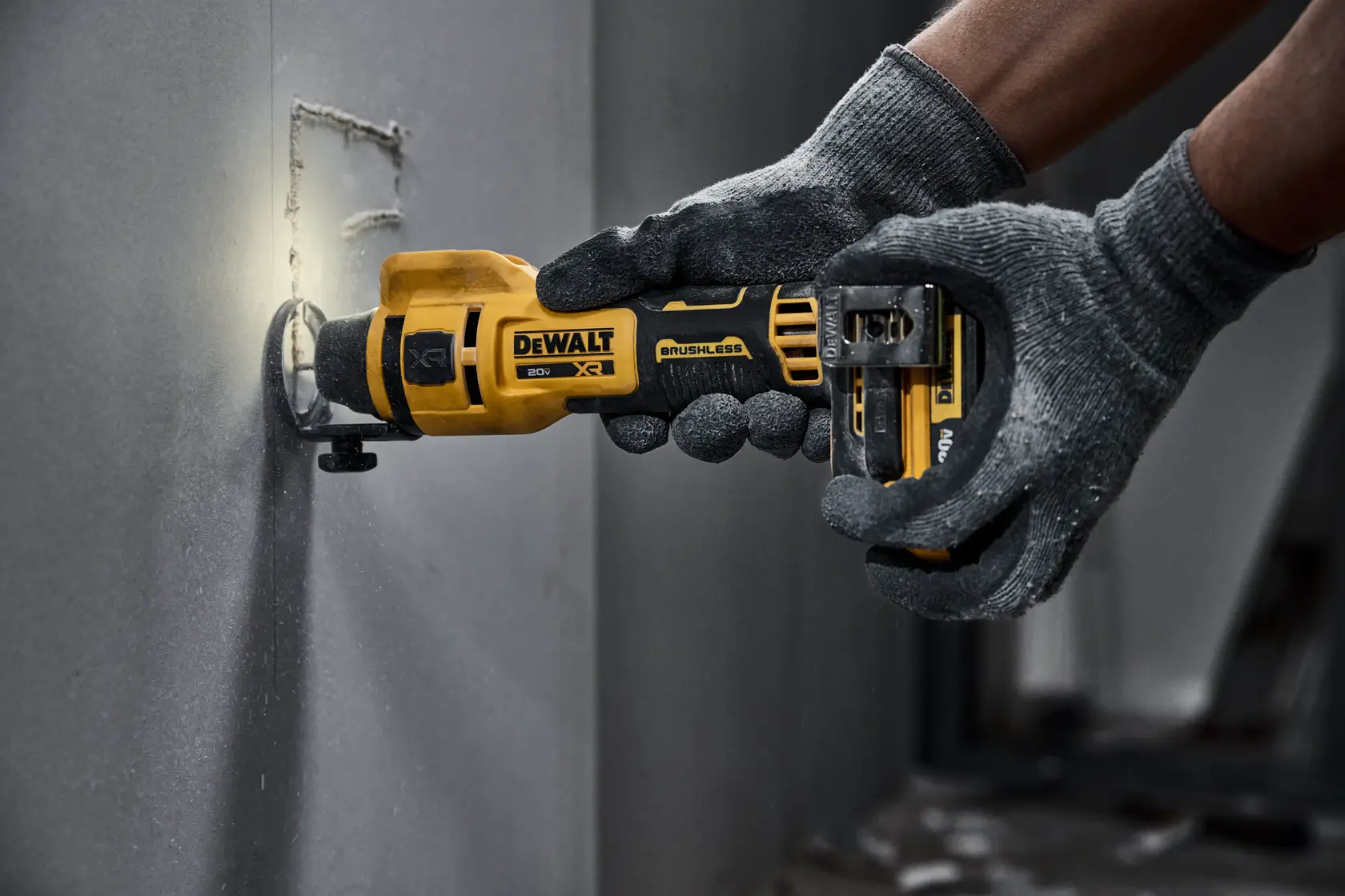 A person wearing gray gloves holds a DEWALT cordless rotary tool and uses it to cut a hole in a gray wall.