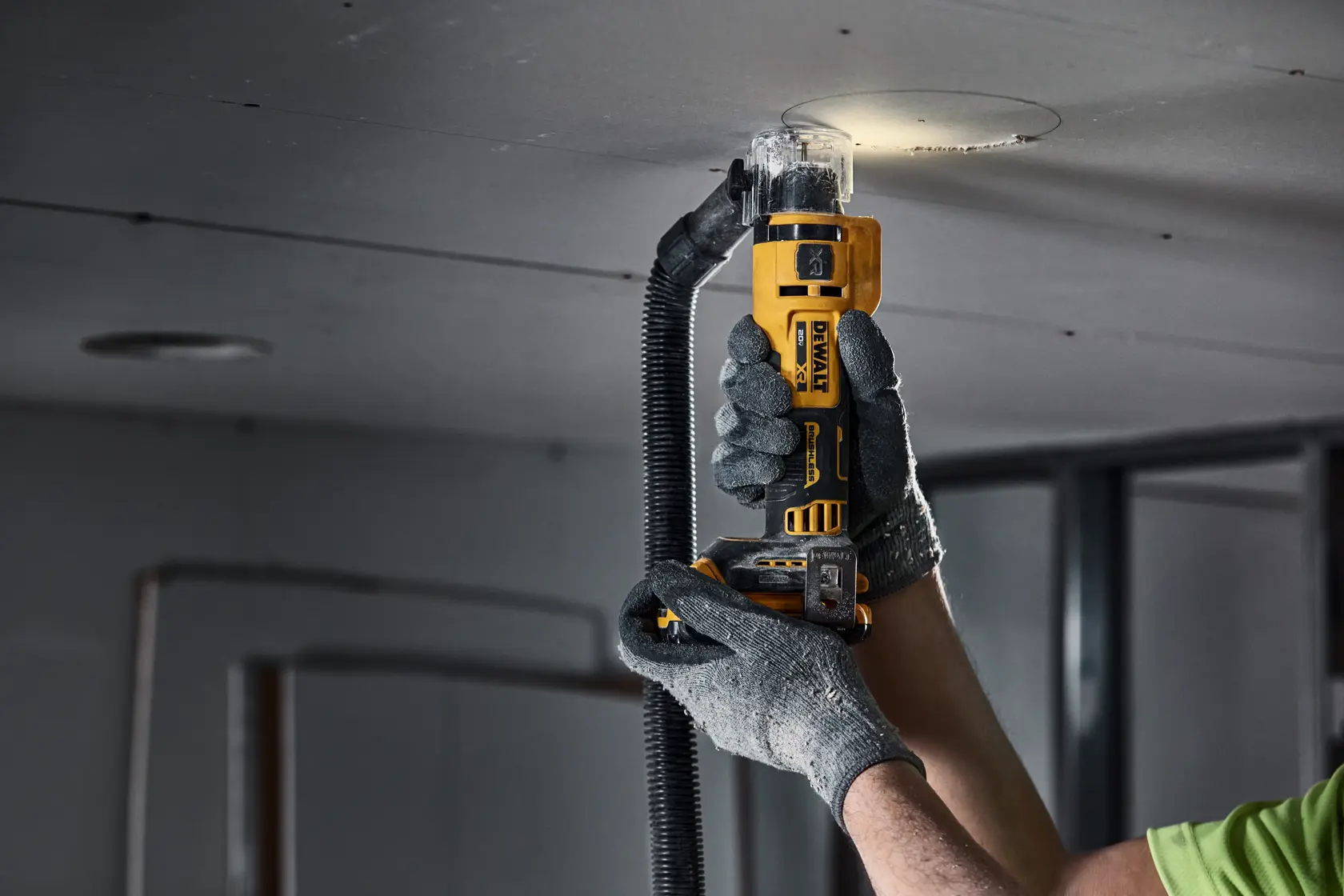A person wearing work gloves uses a DEWALT rotary tool to cut a circular opening in a ceiling panel. The tool is attached to a dust extraction hose and emits light at the cutting area.