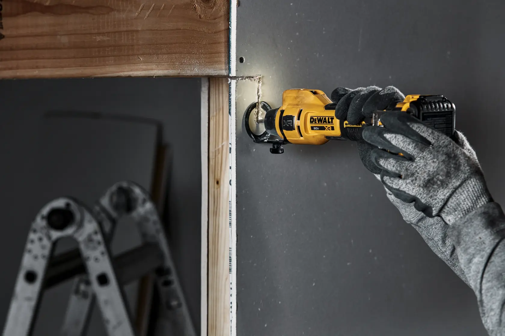 A person wearing gray gloves is using a yellow DEWALT cordless rotary tool to cut into drywall near a wooden frame. There is a metal ladder in the background.