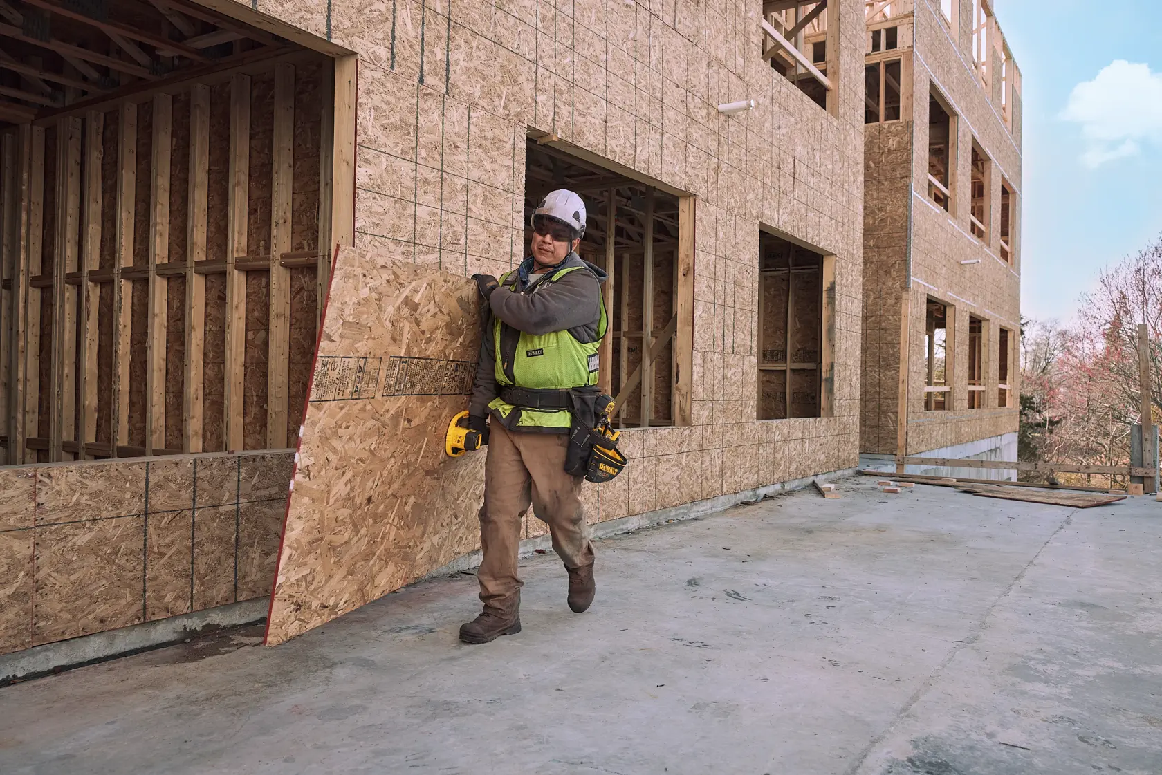 A construction worker wearing a safety vest and helmet is carrying a large sheet of plywood outside a building under construction. The worker has a tool belt and a yellow DEWALT power tool attached to their waist.
