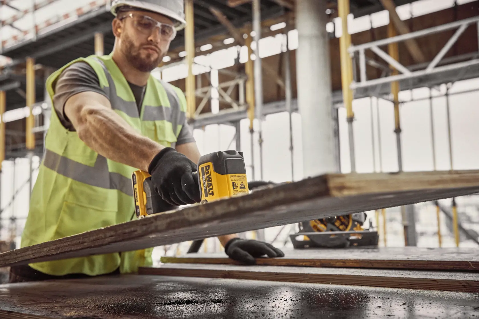 A construction worker wearing a safety vest and gloves is using a DEWALT DCE592D1 photography product on a wooden plank at a building site, with scaffolding visible in the background.