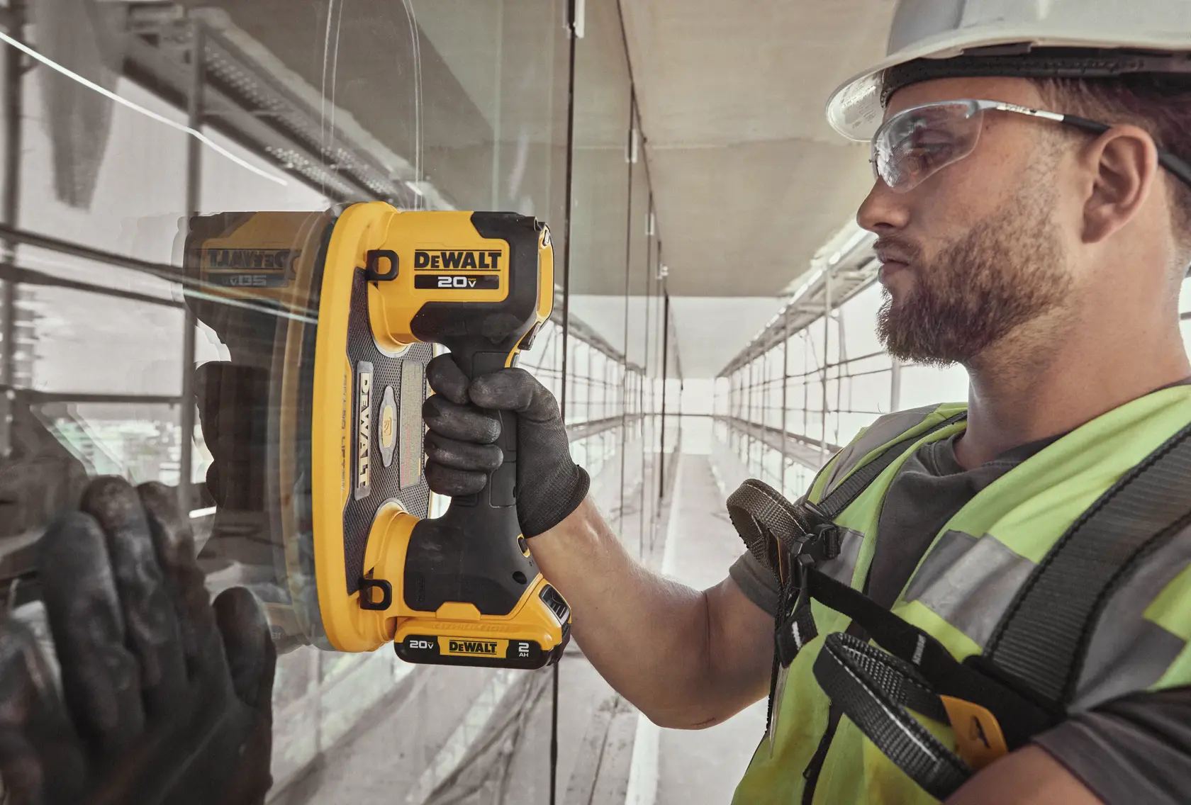 A person wearing safety gear uses a DEWALT 20V battery-powered suction cup tool to lift or install a glass panel in a construction or industrial setting. The person's face is blurred for privacy.