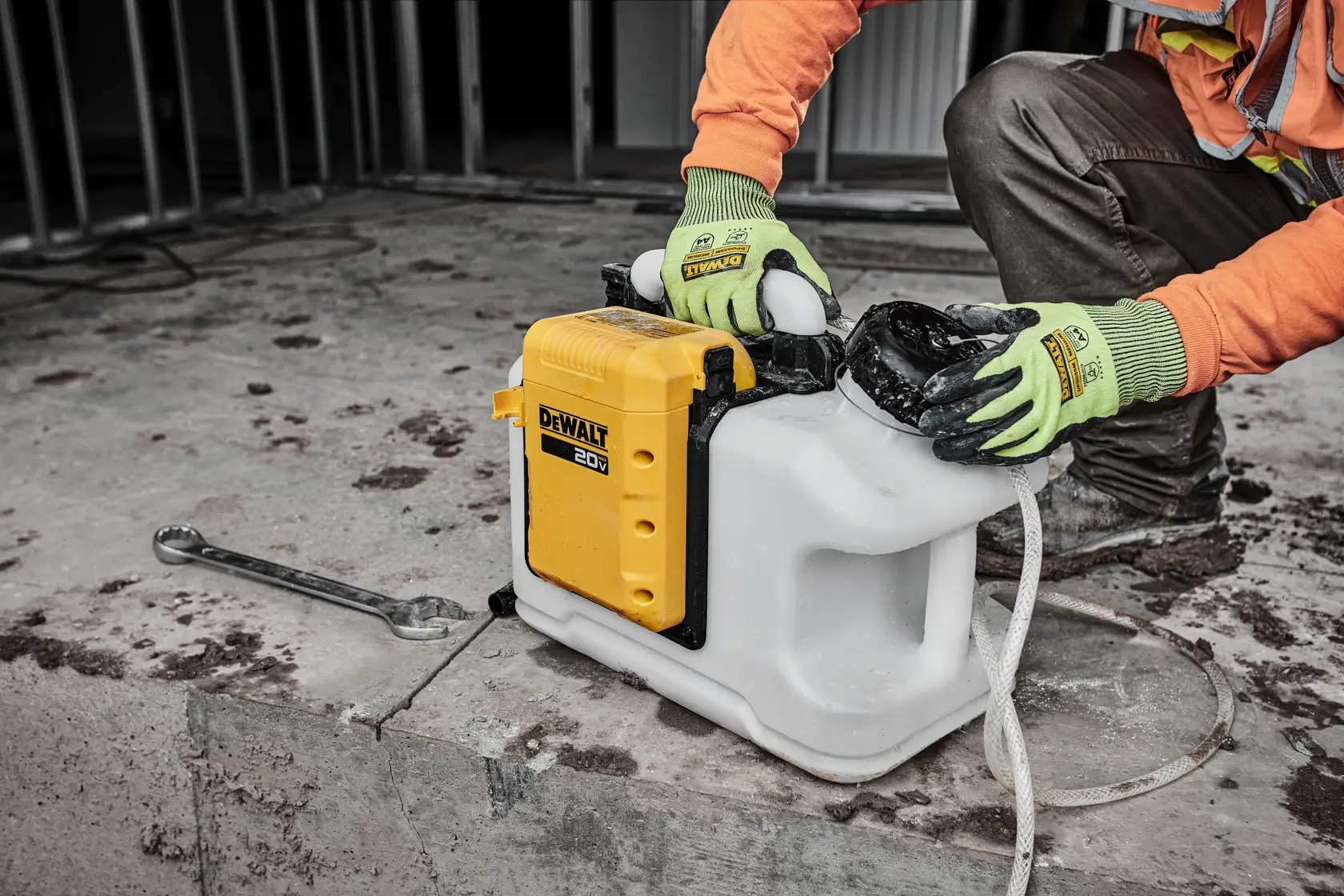 A person wearing work gloves and an orange long-sleeve shirt is handling a DEWALT 20V cordless concrete vibrator placed on a construction site floor, with a wrench lying nearby.