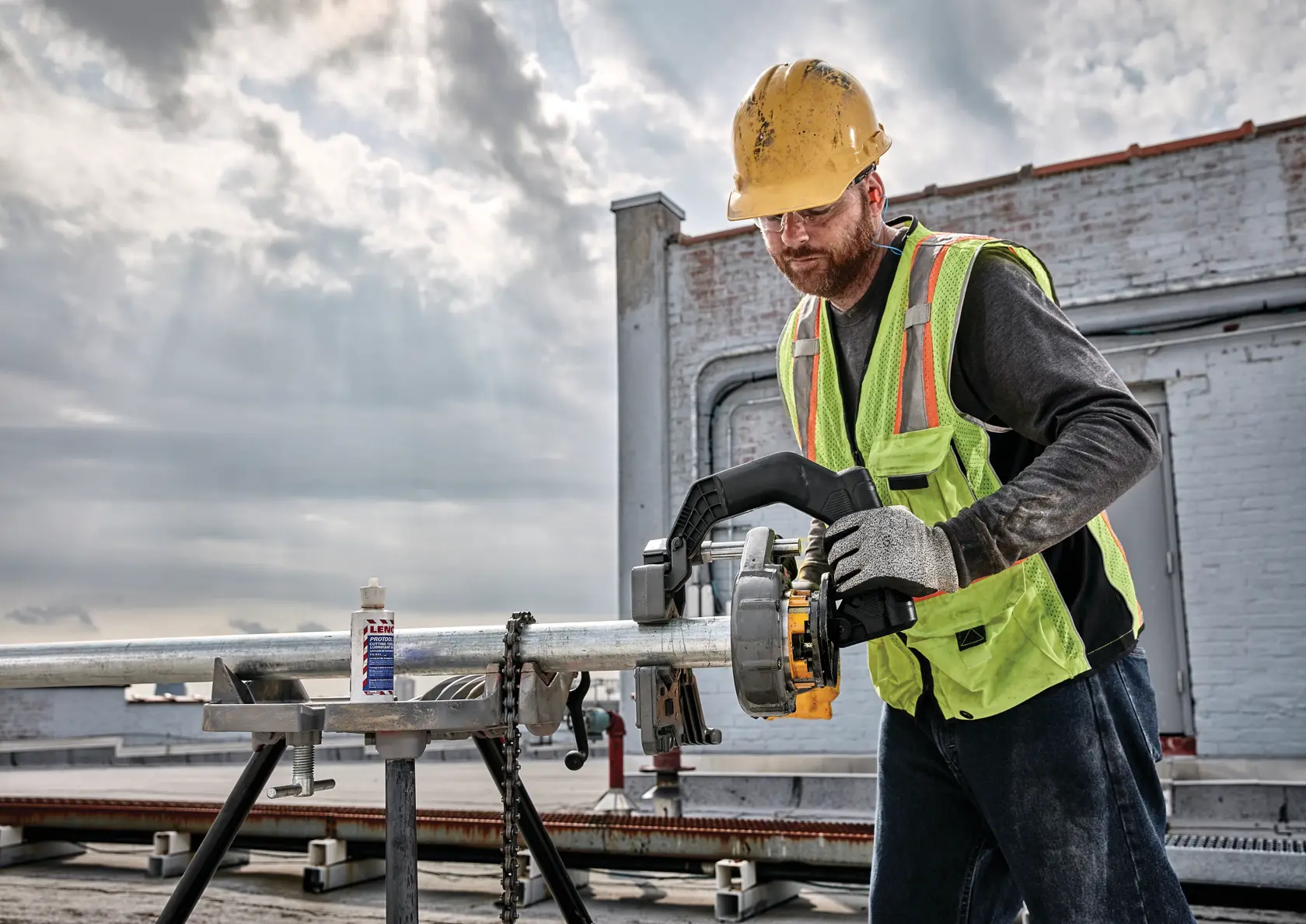 A construction worker wearing a yellow safety helmet and a neon safety vest is using a DEWALT pipe threader to work on a metal pipe outdoors. The worker is also wearing gloves, and there is a bottle of lubricant on the workbench. The scene takes place on a rooftop with a cloudy sky in the background.
