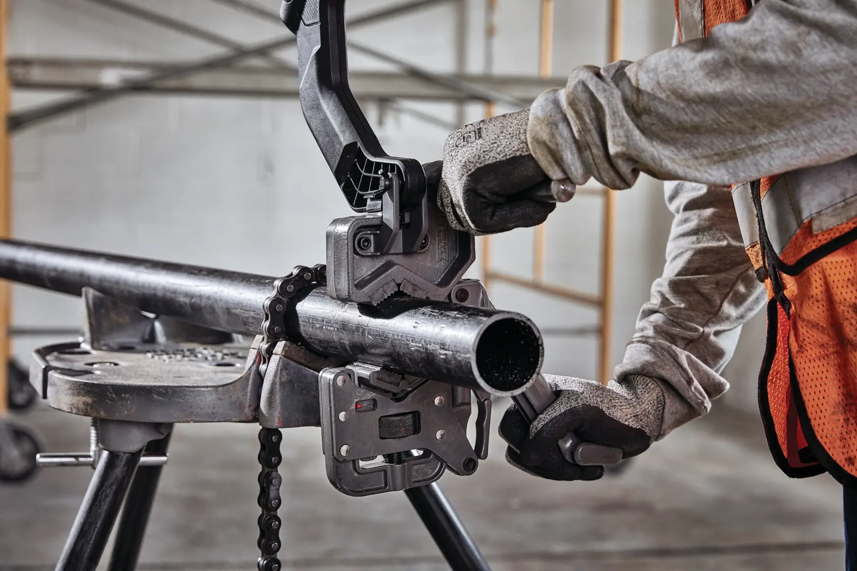 Person wearing safety gloves and an orange vest using a DEWALT pipe threading tool to work on a metal pipe in an industrial indoor setting.
