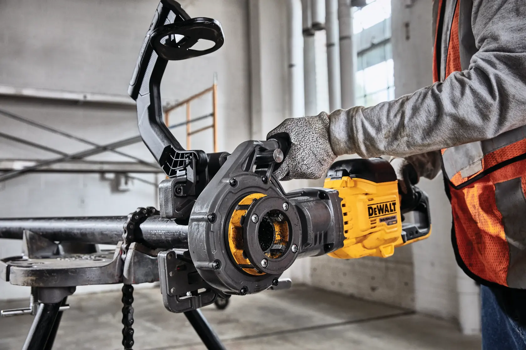 A worker is using a DEWALT DCE700X2 pipe threading machine to thread a metal pipe in an industrial setting. The worker is wearing gloves and a safety vest.