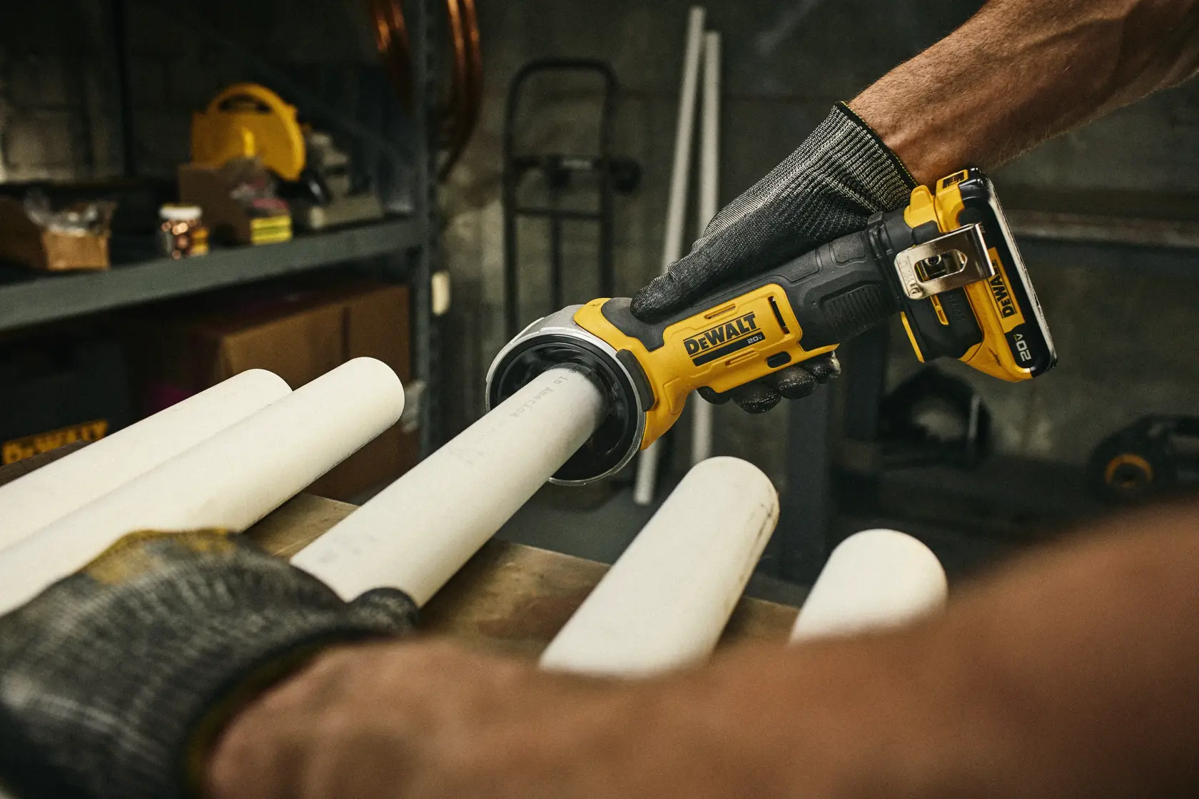 A person wearing gloves is using a yellow and black DEWALT cordless pipe cutter to cut a white PVC pipe in a workshop setting. Several pipes are placed on the workbench and there are tools and equipment visible in the background.