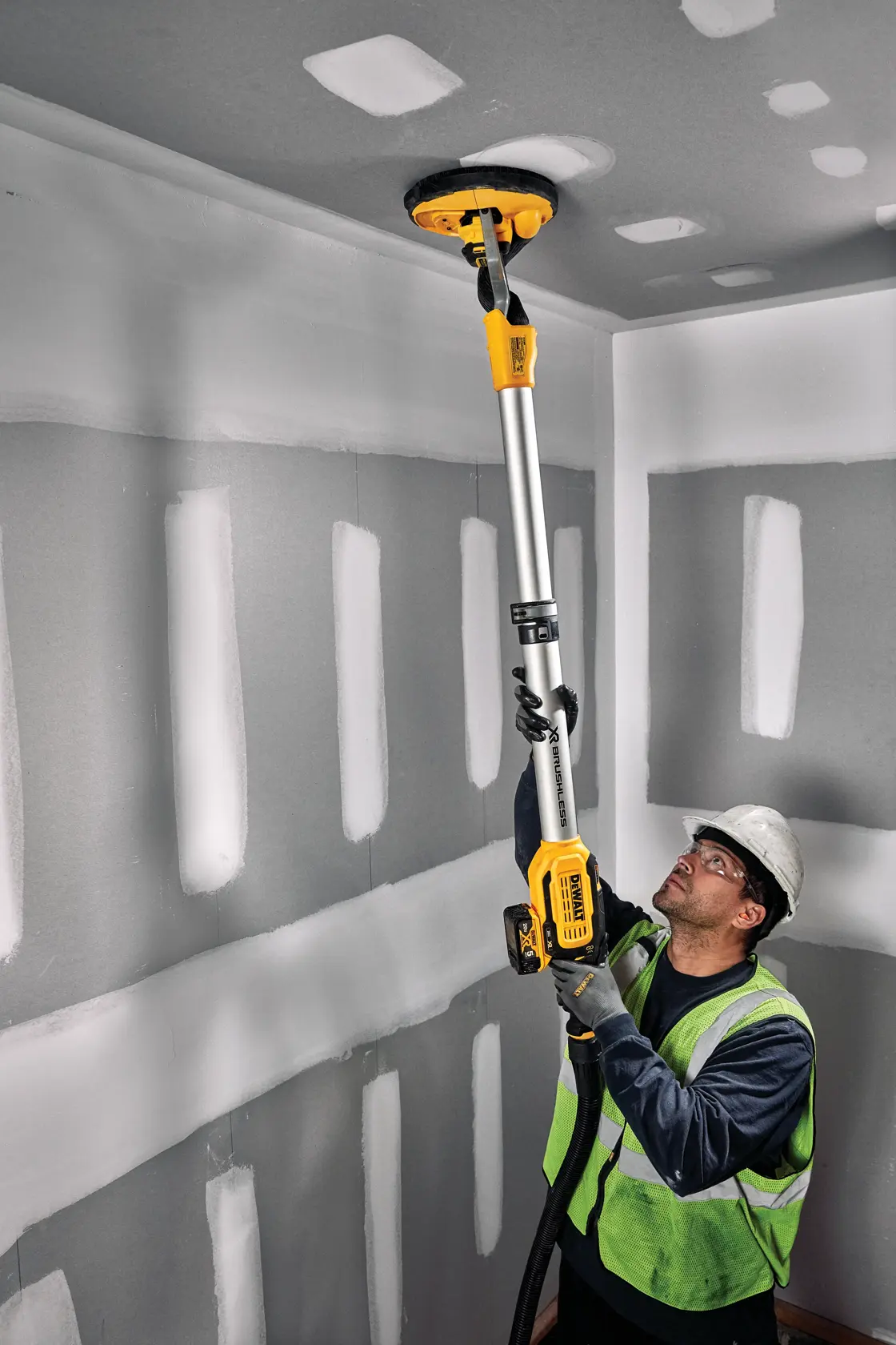 A person wearing a safety vest and helmet uses a DEWALT DCE800P2 drywall sander to sand the ceiling in a room with unfinished drywall.