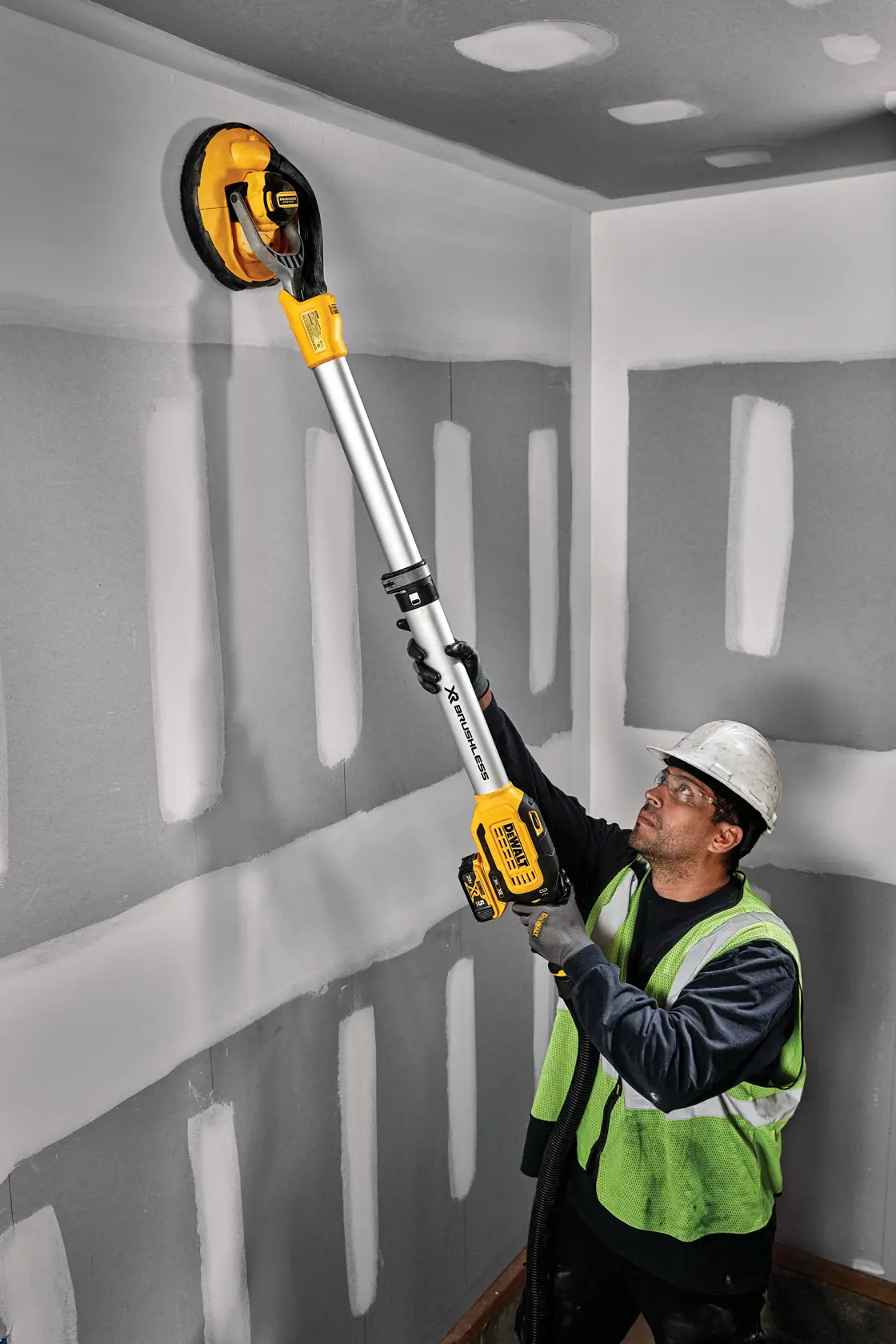A worker wearing a safety vest and helmet uses a DEWALT DCE800P2 cordless drywall sander to sand a wall and ceiling in a room under construction.