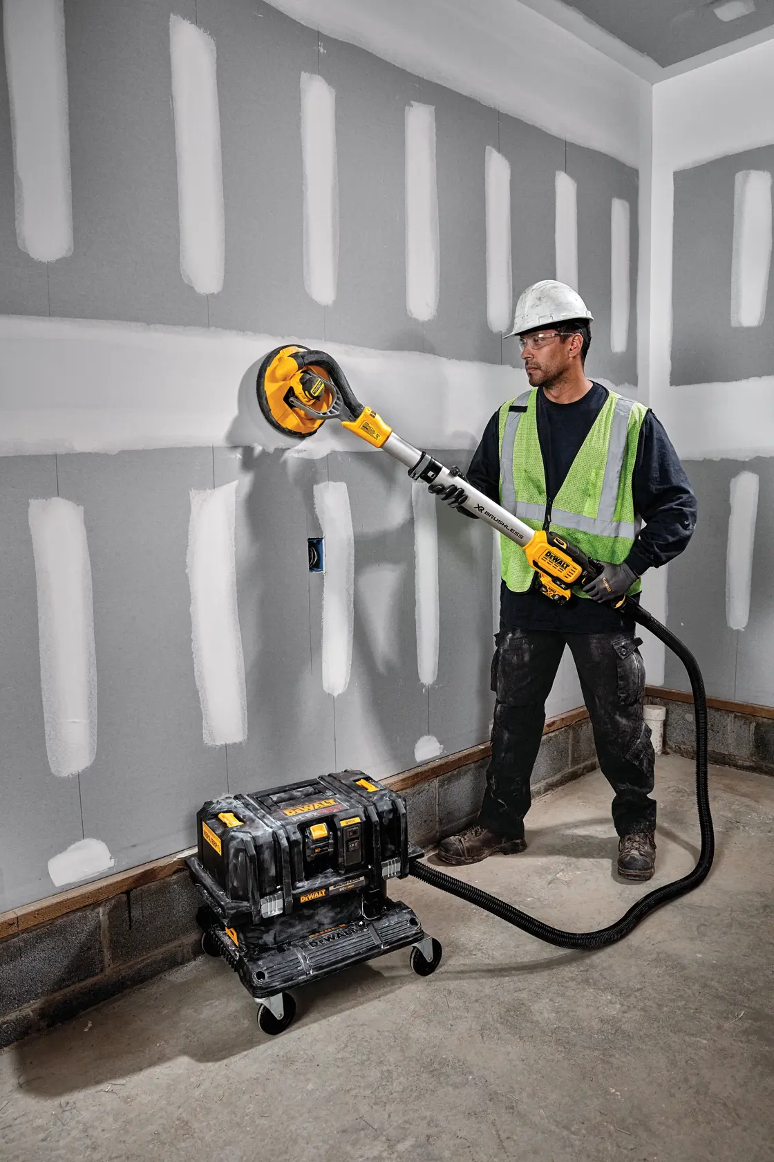 A worker in a safety vest and hard hat using a DEWALT drywall sander with a vacuum attachment, sanding a wall in a construction environment. The vacuum equipment is connected to the sander and sits on the floor.
