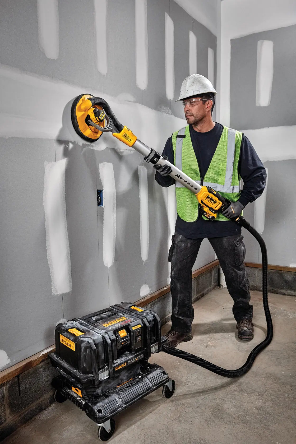 A construction worker wearing a safety helmet and high-visibility vest uses a DEWALT drywall sander connected to a portable dust extraction system. The worker is sanding drywall in a room with unfinished walls.