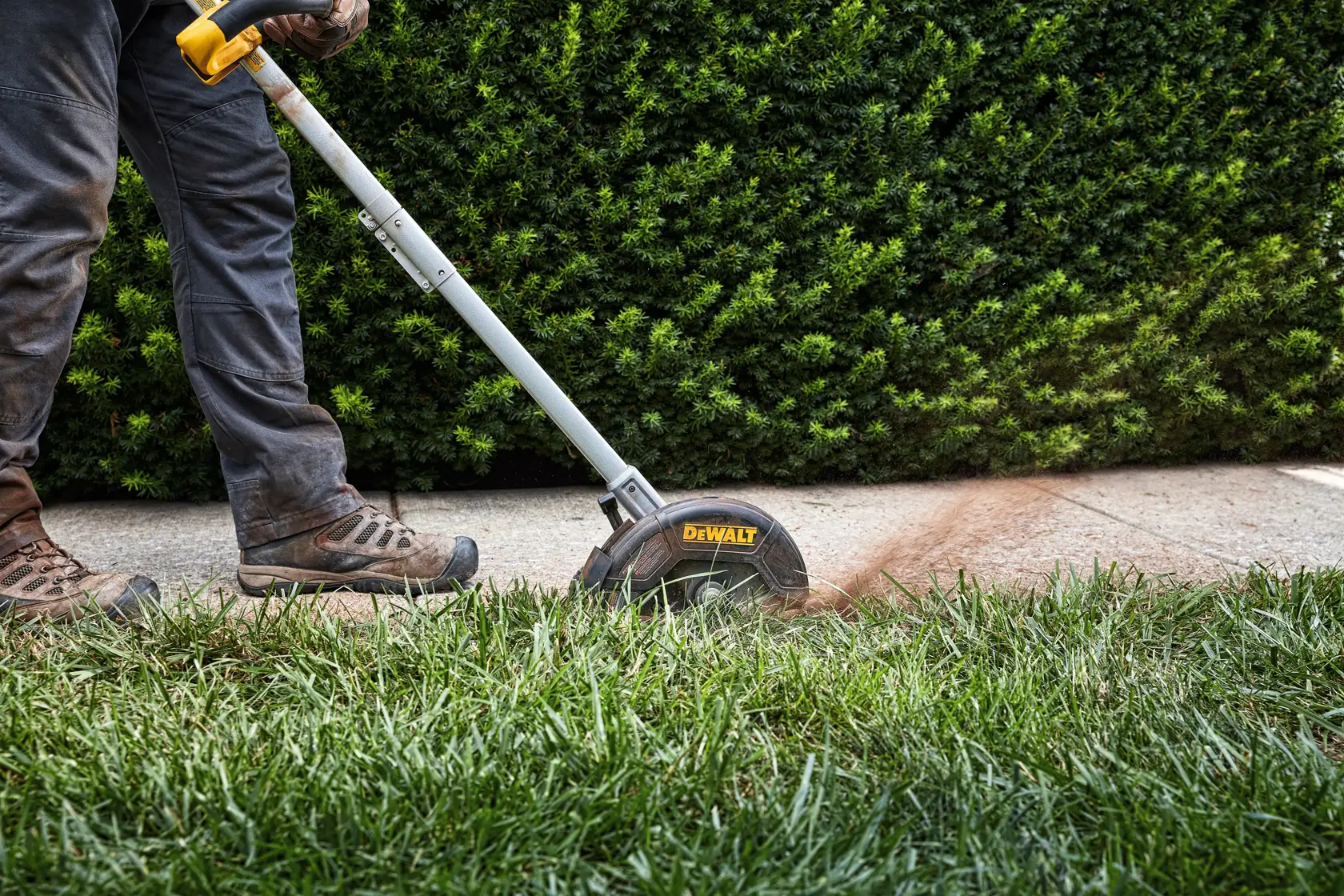 A person uses a DEWALT cordless edger to trim the edge of a lawn next to a sidewalk, with grass and dirt visibly being cut.