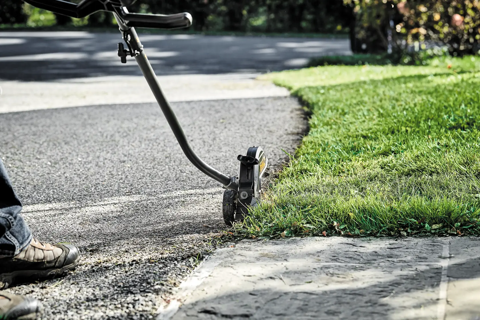 A person is using a DEWALT outdoor power tool to edge the lawn along a sidewalk and driveway, creating a neat boundary between the grass and pavement.