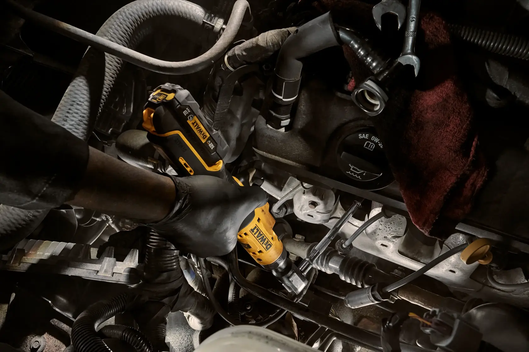 A person wearing black gloves is using a DEWALT cordless ratchet tool inside an engine compartment, surrounded by various automotive parts and hoses.