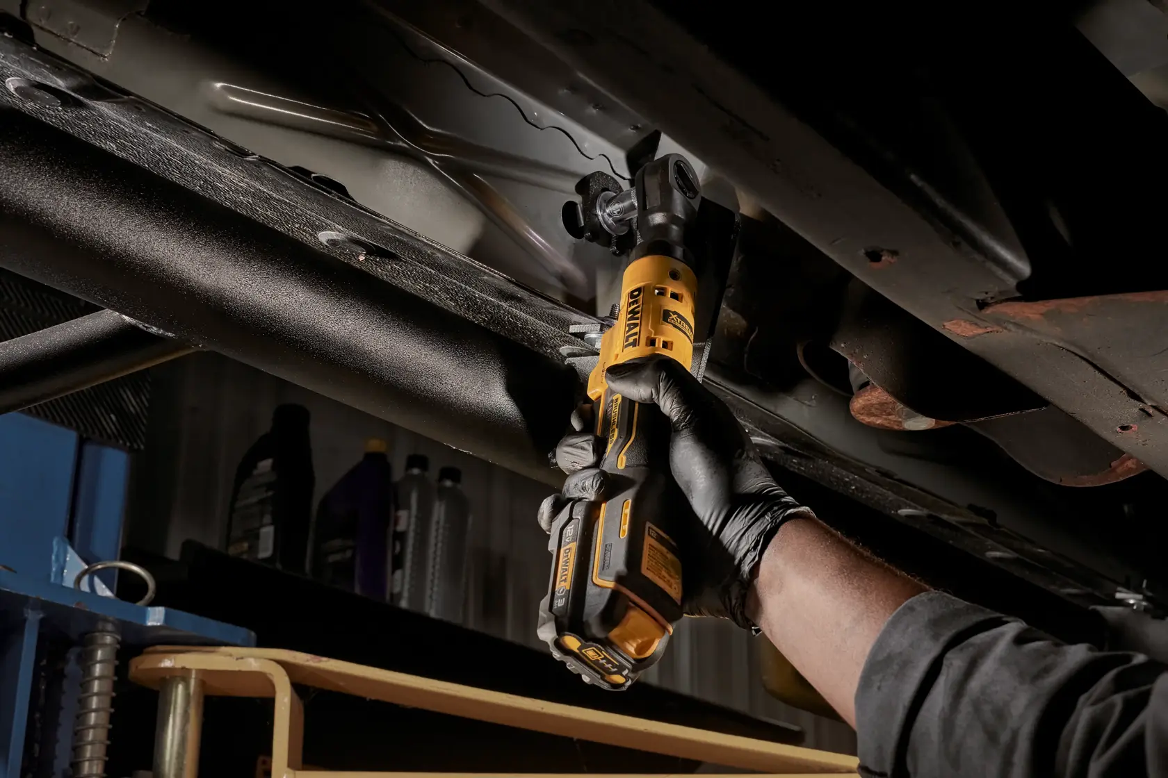 A person wearing black gloves uses a DEWALT cordless ratchet tool to work on a metal structure under a vehicle.