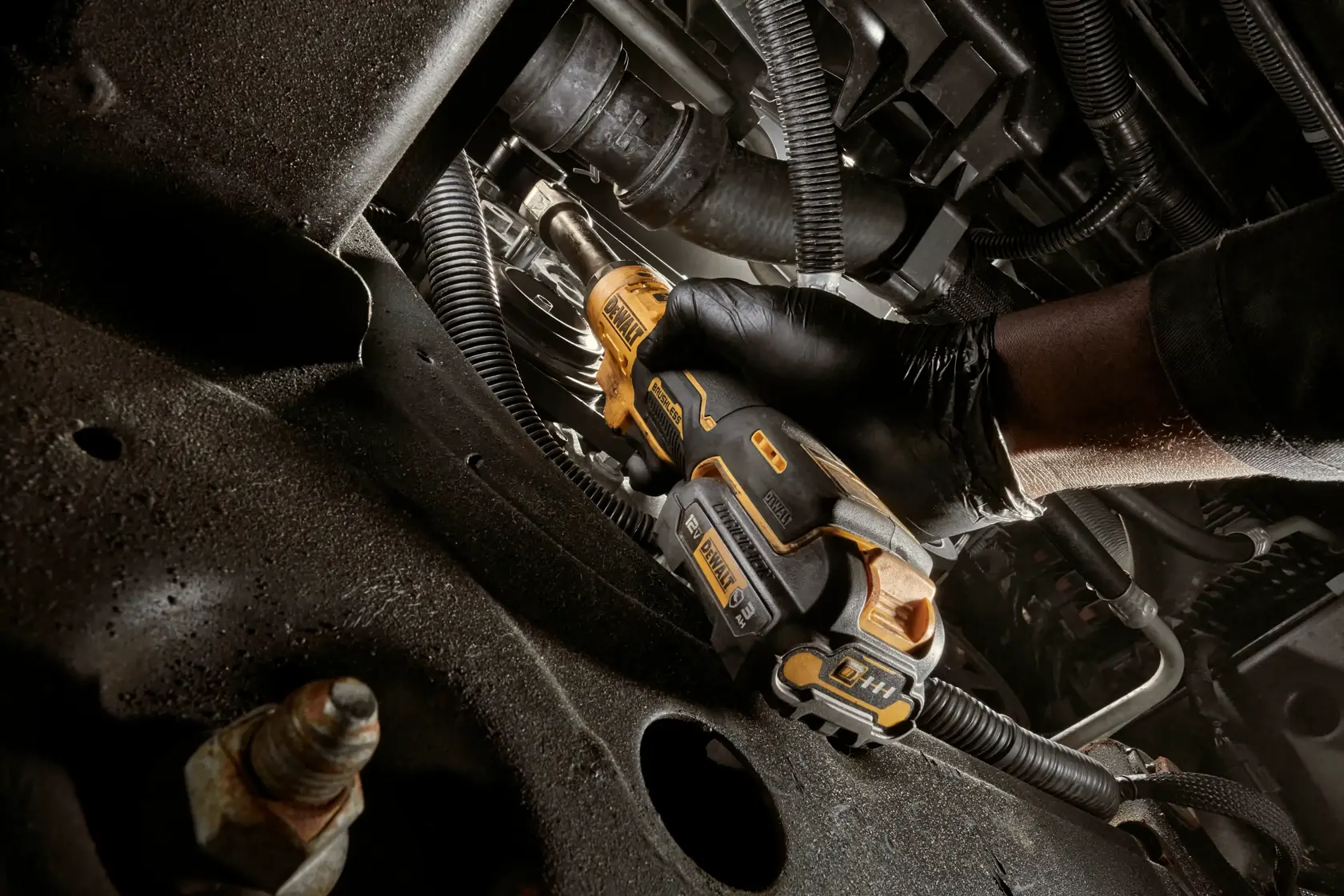 A person wearing black gloves uses a DEWALT cordless electric screwdriver to work on machinery or an automotive engine. The tool is yellow and black, and the environment consists of metal parts and cables.