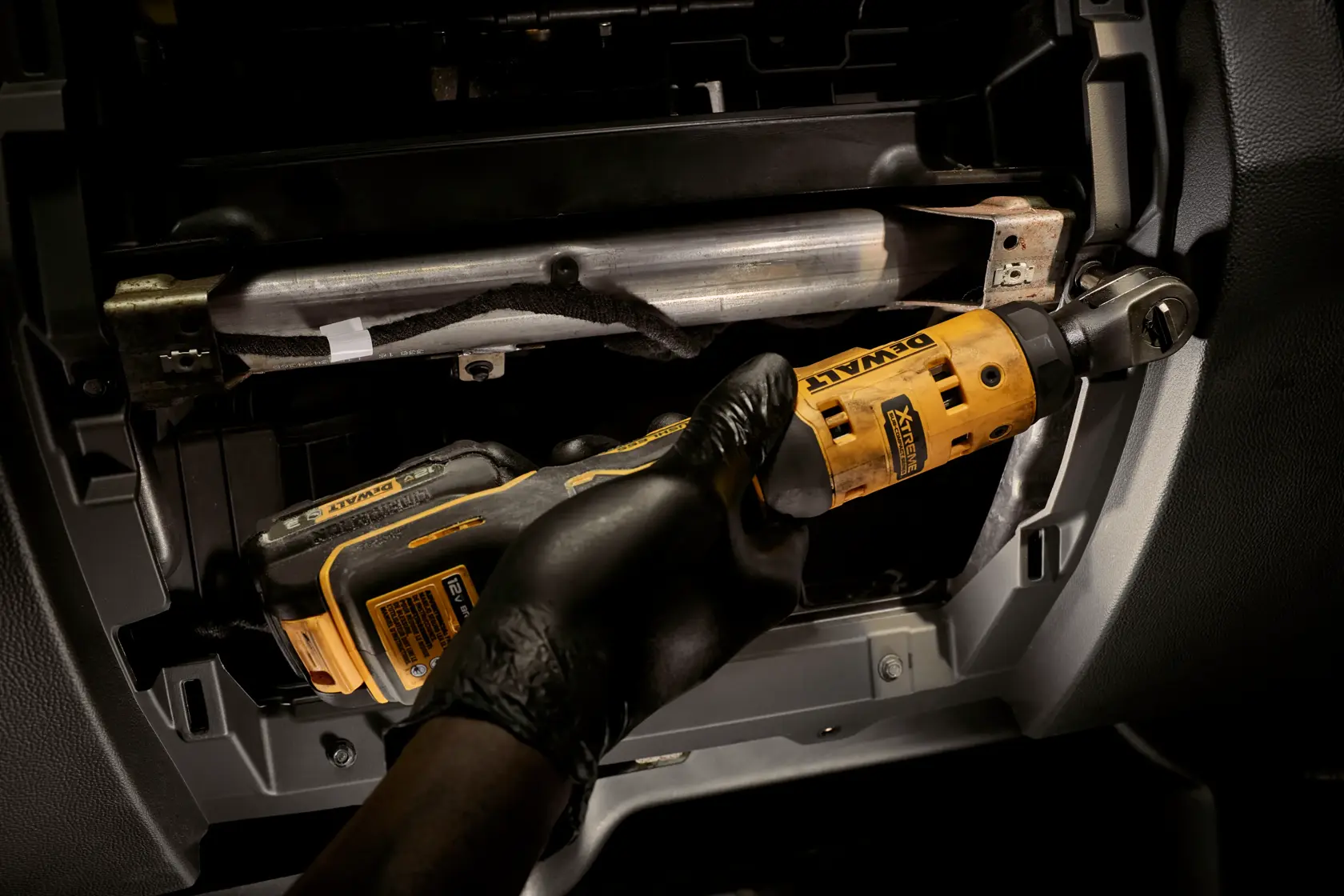 A person wearing a black glove is using a DEWALT cordless ratchet tool inside a mechanical component or vehicle dashboard.
