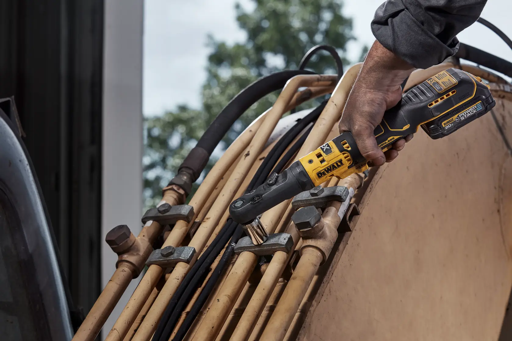 A person using a DEWALT cordless ratchet tool to tighten or loosen fittings on a set of industrial pipes outdoors.