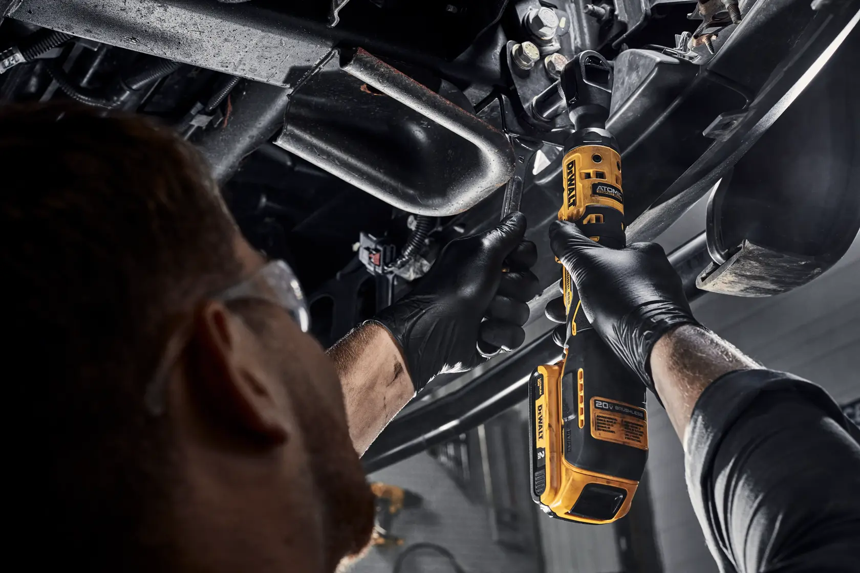 A person wearing safety glasses and black gloves uses a yellow DEWALT cordless ratchet tool to work on mechanical parts underneath a vehicle.