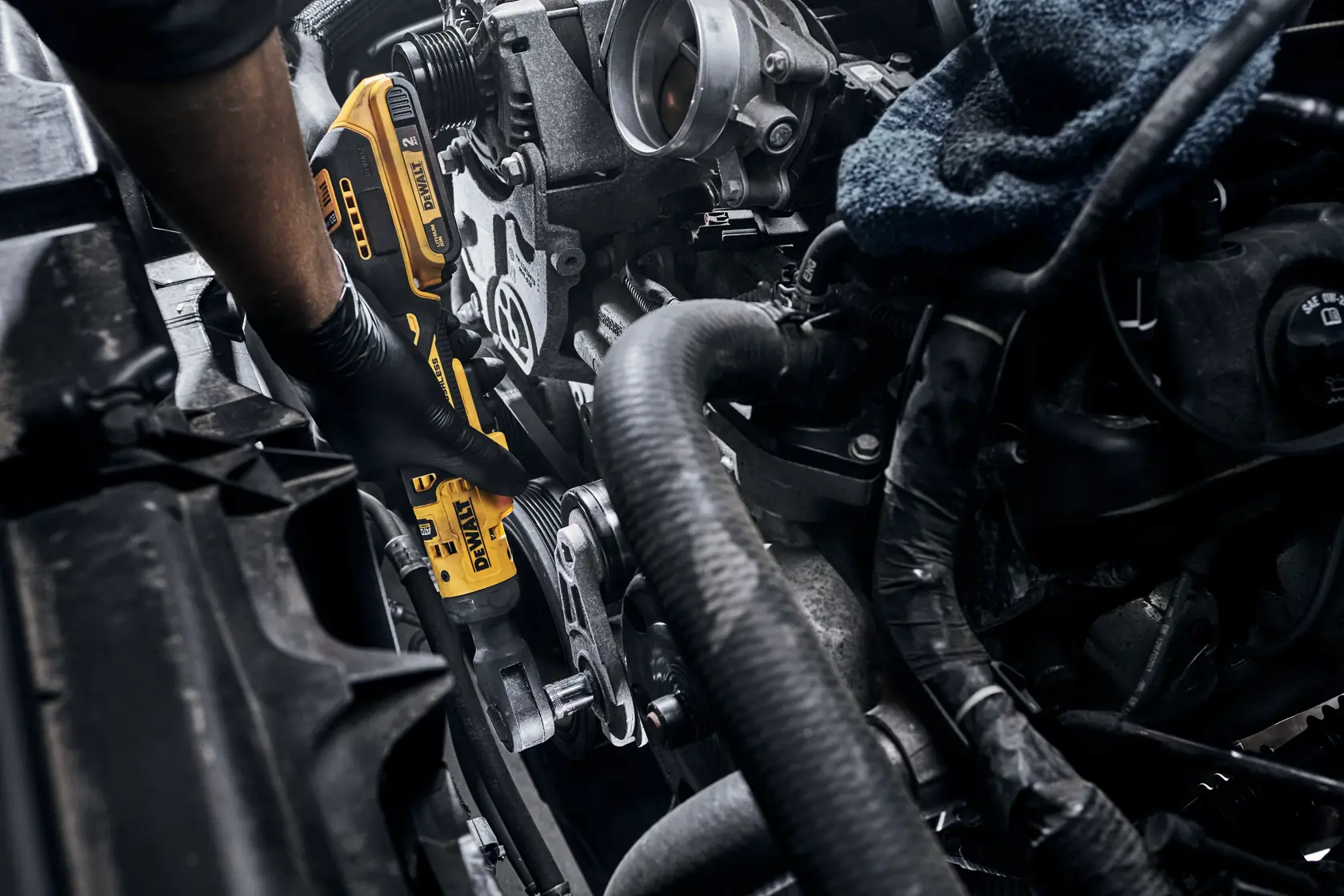 A person wearing a black glove is using a yellow DEWALT cordless ratchet tool to work on a car engine.