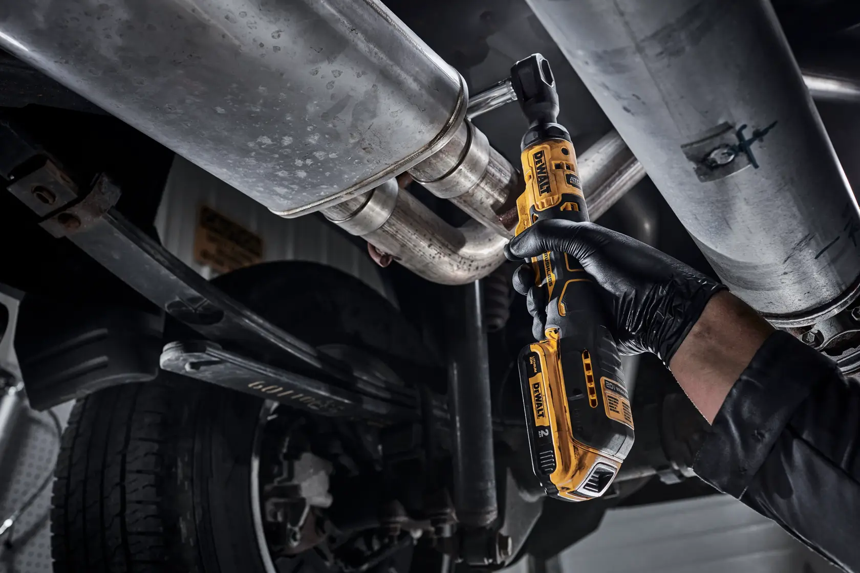 A person wearing a black glove uses a DEWALT cordless ratchet wrench to work on the underside of a vehicle, fastening a bolt on a metal exhaust pipe.