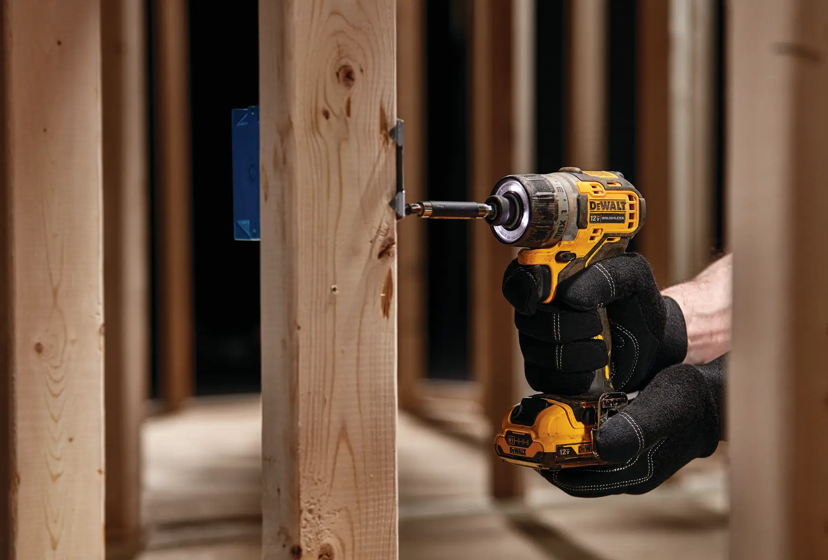 A person wearing black gloves uses a yellow DEWALT cordless drill to drive a screw into a wooden beam in a construction setting.