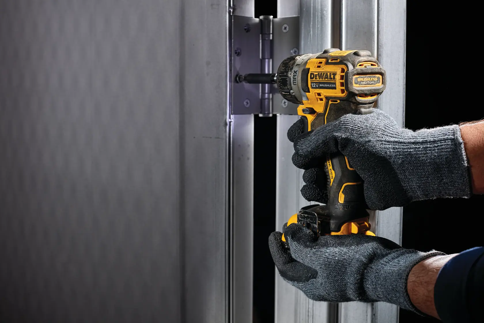 A person wearing grey gloves is using a yellow DEWALT cordless drill to install or tighten screws on a metal hinge attached to a metallic door frame.