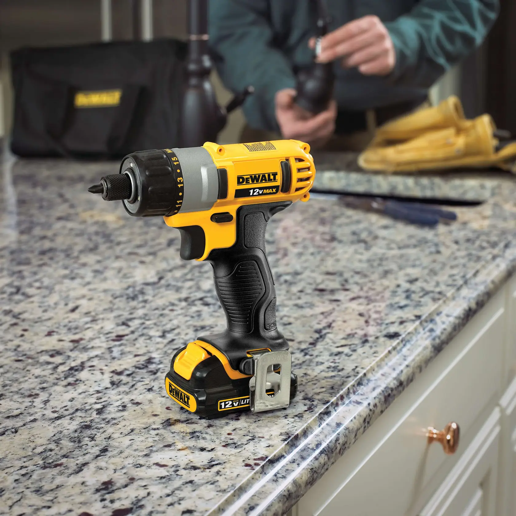 A DEWALT 12V MAX cordless drill with battery, standing upright on a granite countertop in a kitchen. In the background, a person is working with some tools and gloves.