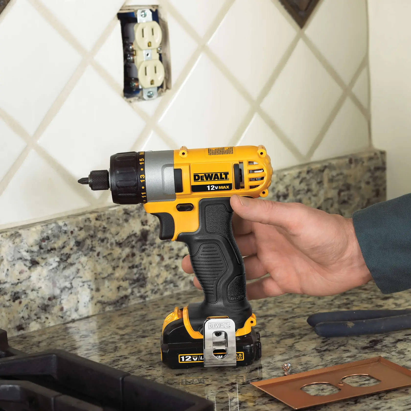A person holding a yellow and black DeWalt 12V MAX cordless drill on a kitchen countertop, with a granite surface and tile backsplash. The drill is near an electrical outlet being worked on and some tools and outlet covers are visible nearby.
