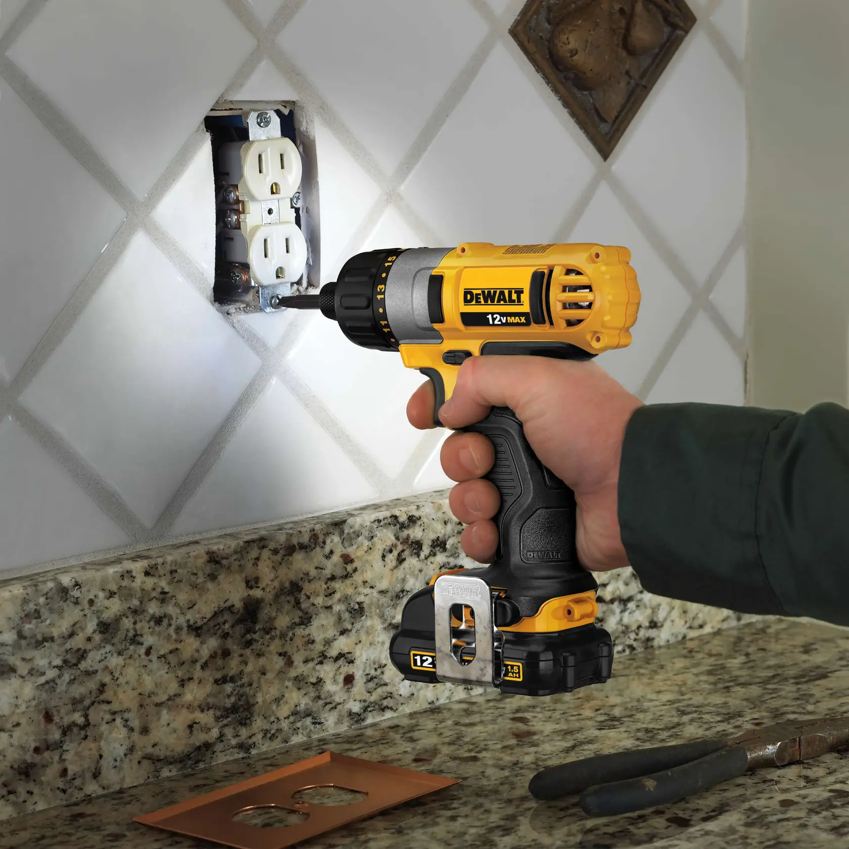 A person using a yellow DEWALT 12V MAX cordless drill to install or remove screws from a wall electrical outlet, with the drill positioned near a tiled backsplash and a granite countertop.