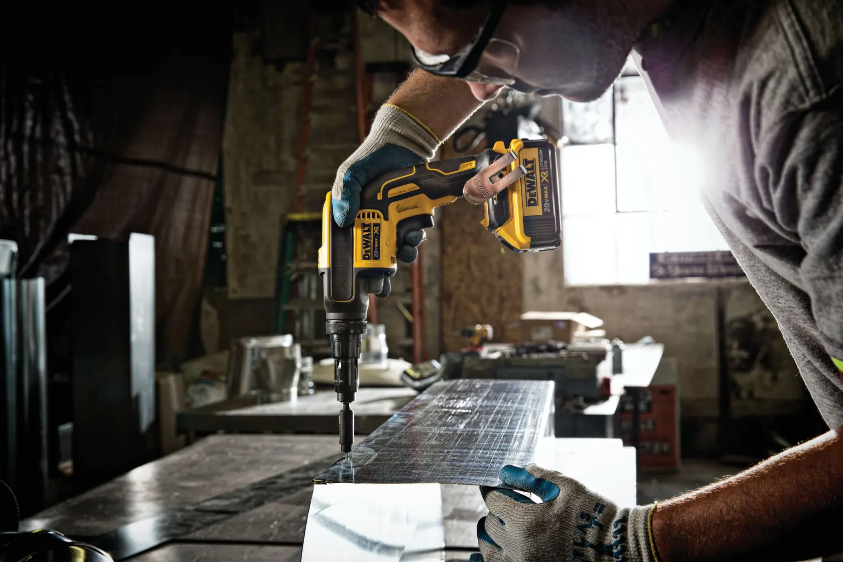 A person wearing gloves and safety glasses uses a DEWALT cordless power tool to work on a metal sheet in an industrial workshop.