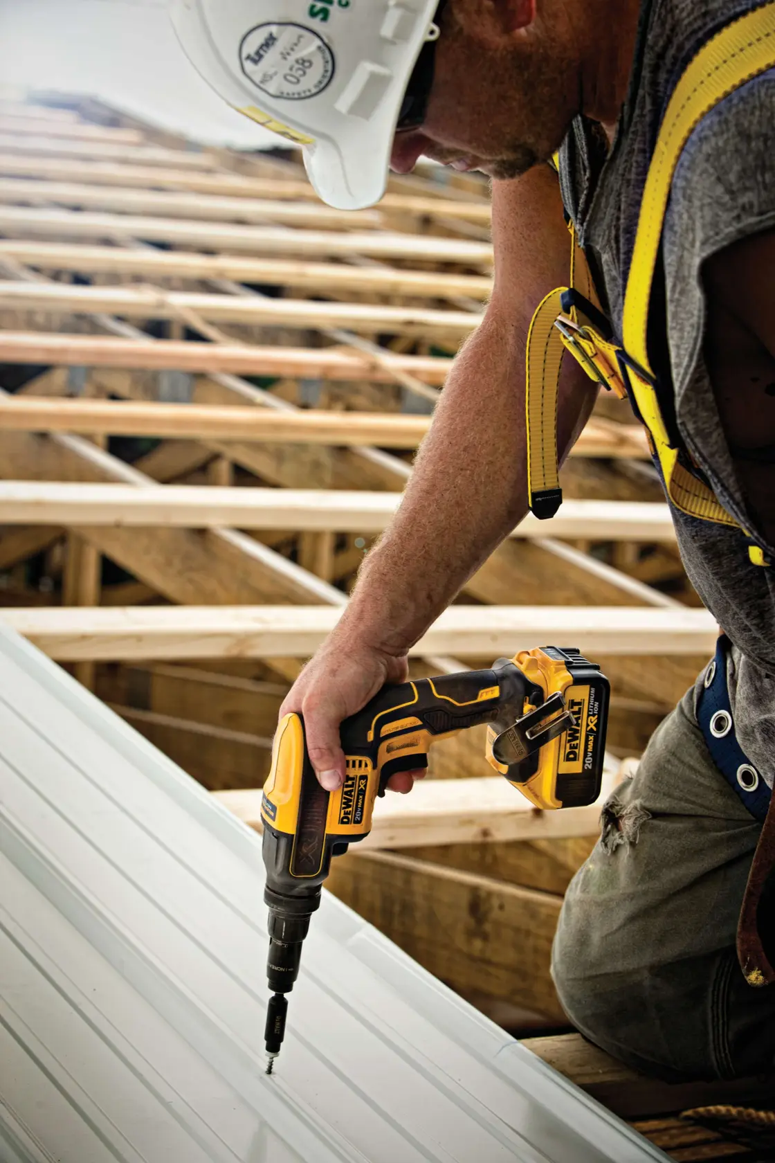 A worker wearing a safety harness and helmet uses a DEWALT cordless screwdriver to fasten a screw into a metal sheet on a construction site.
