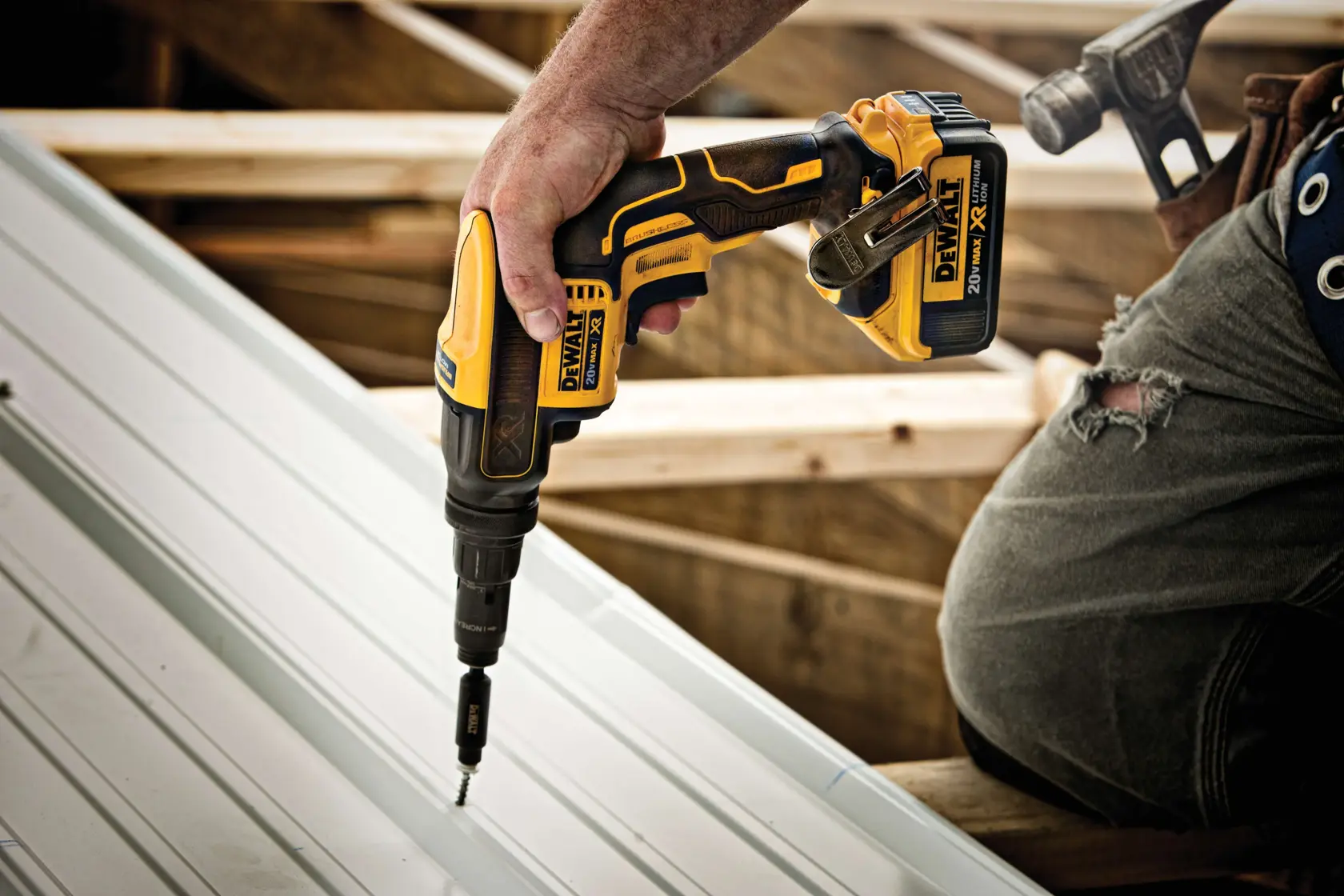 A person using a DEWALT cordless screwdriver to install screws into a metal sheet, with visible construction framing and a hammer attached to their belt.