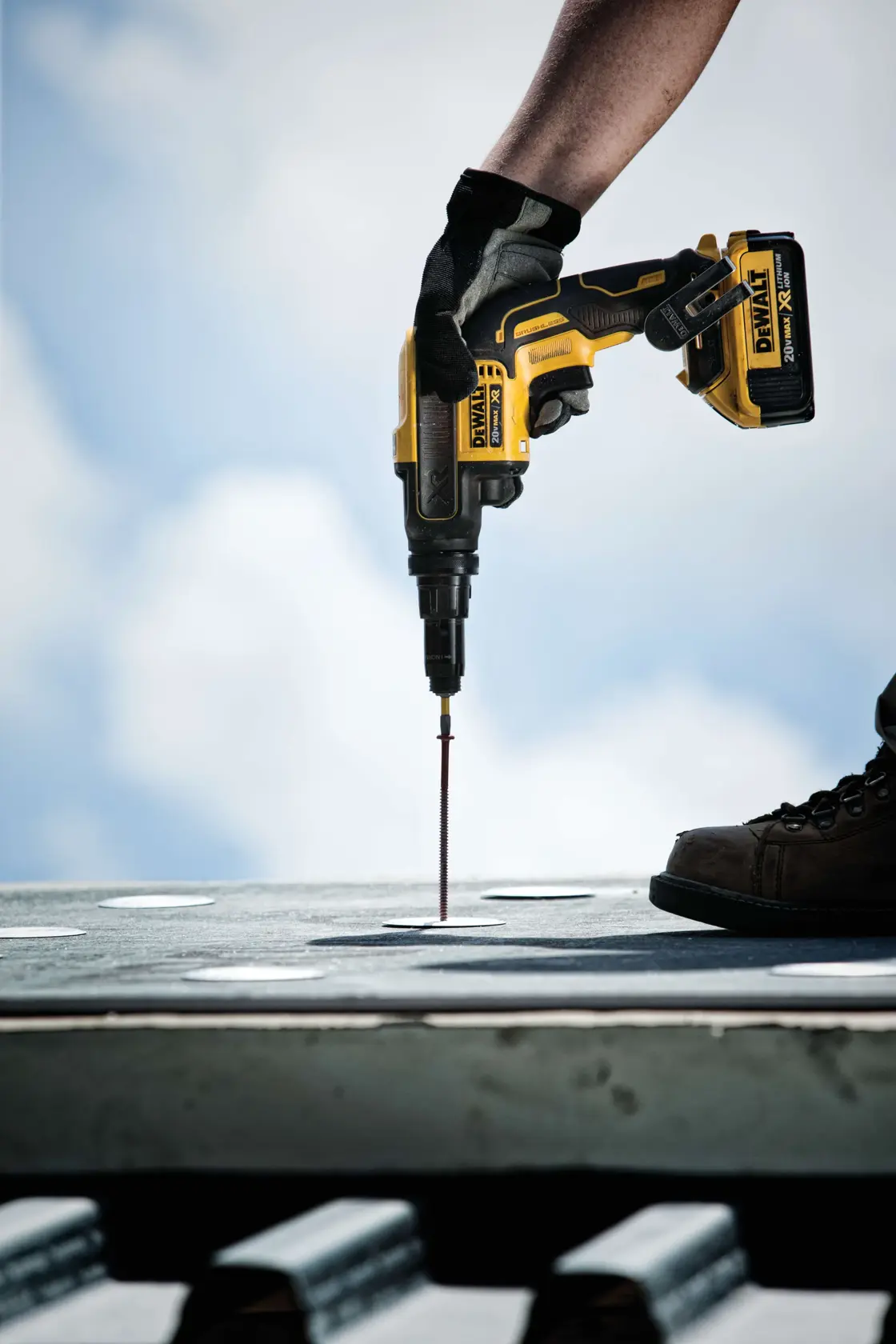 A person wearing work gloves is using a DEWALT cordless power drill to drive a screw into a metal surface, with the sky visible in the background.