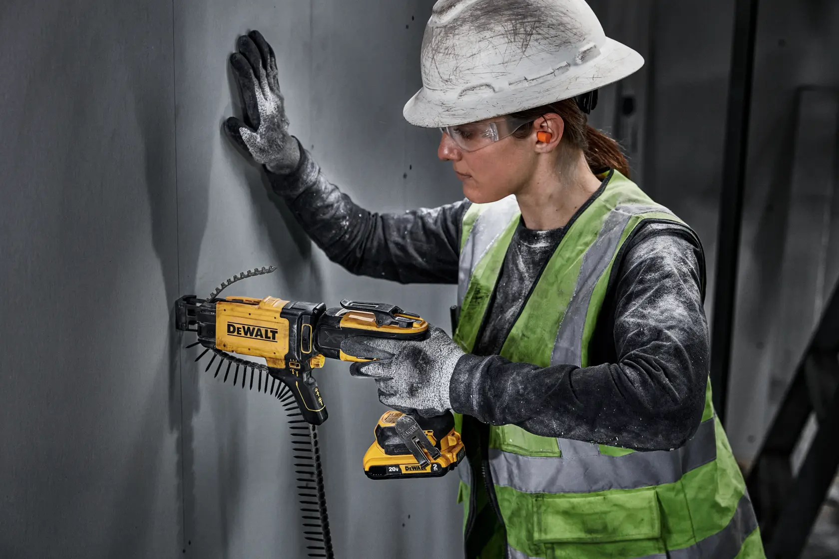 A construction worker wearing a hard hat and a reflective safety vest uses a DEWALT DCF630D2 drywall screw gun to attach panels to a wall. The worker's face is blurred for privacy.