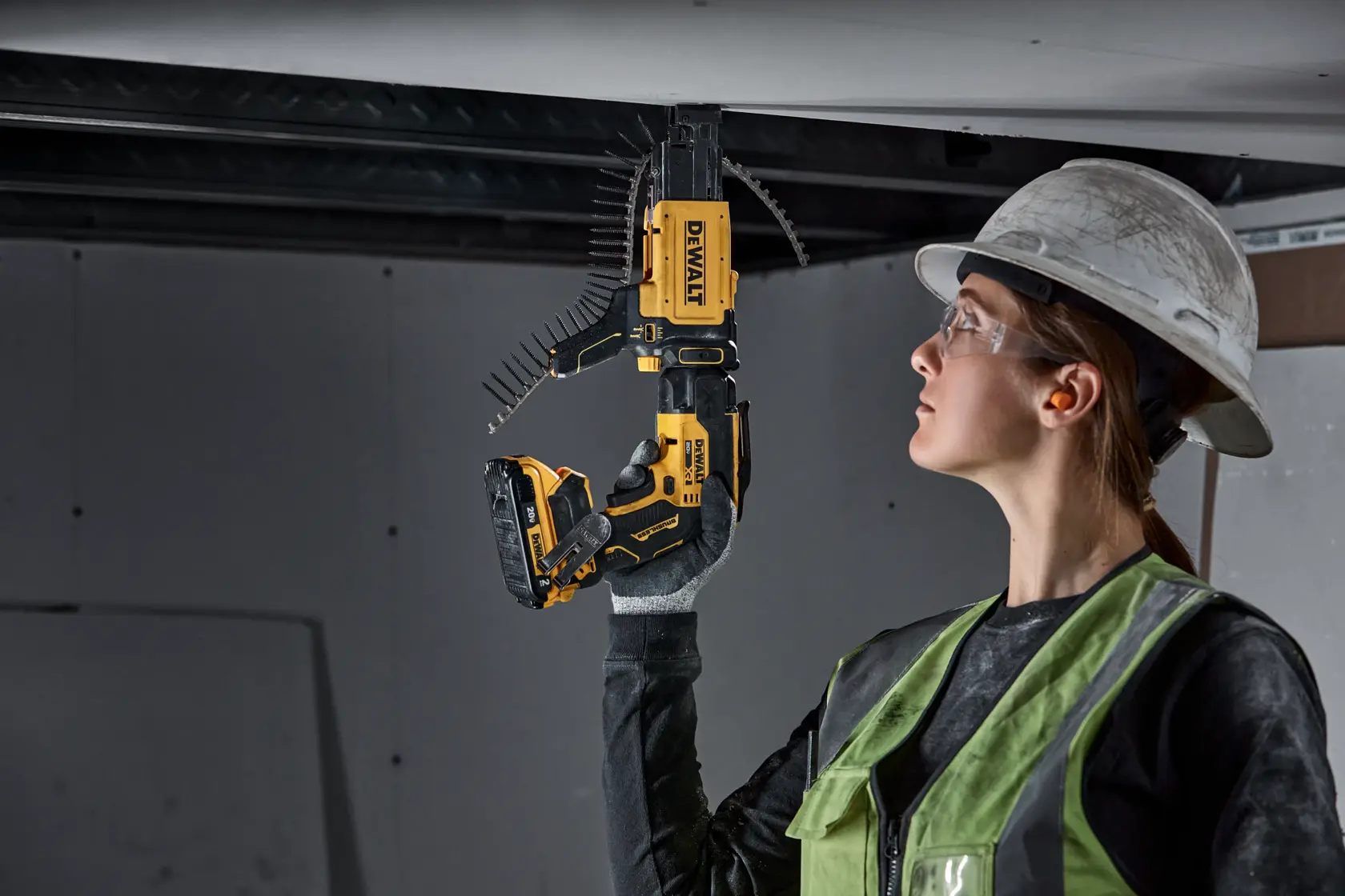A construction worker wearing safety gear uses a DEWALT cordless screw gun (DCF630D2) to install drywall on a ceiling.