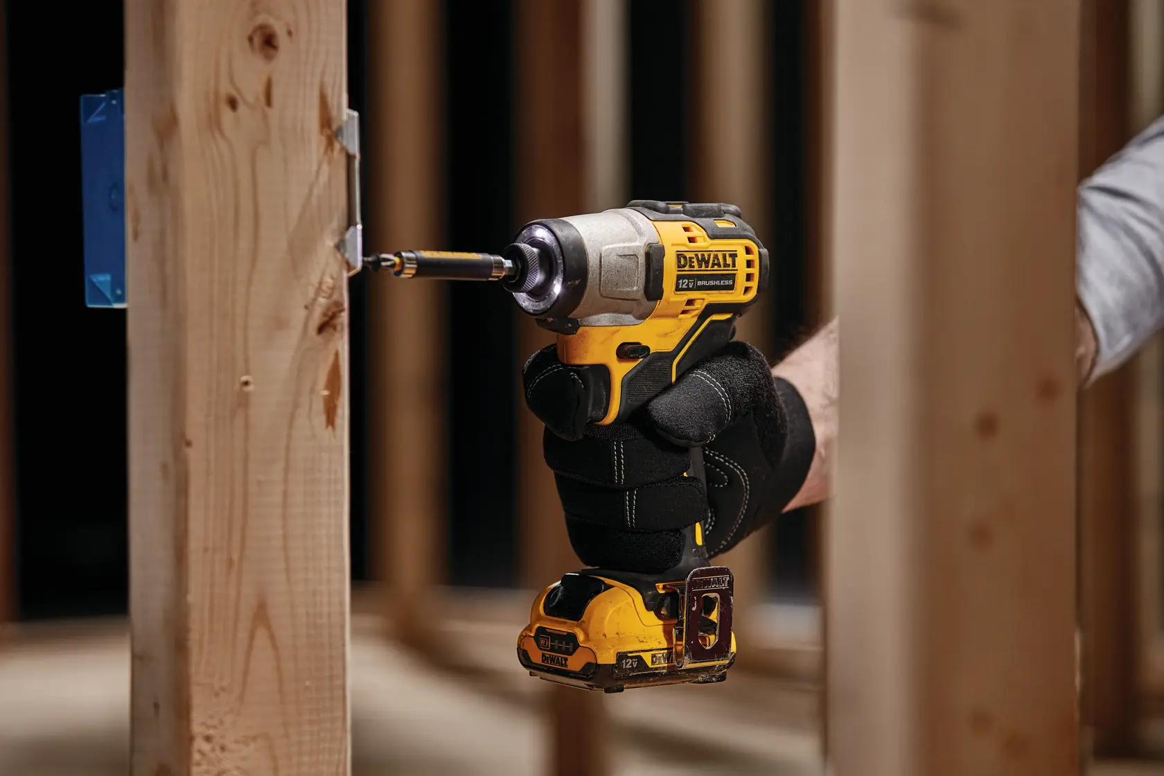 A person wearing black work gloves uses a yellow and black DEWALT cordless impact driver to drill a screw into a wooden frame.
