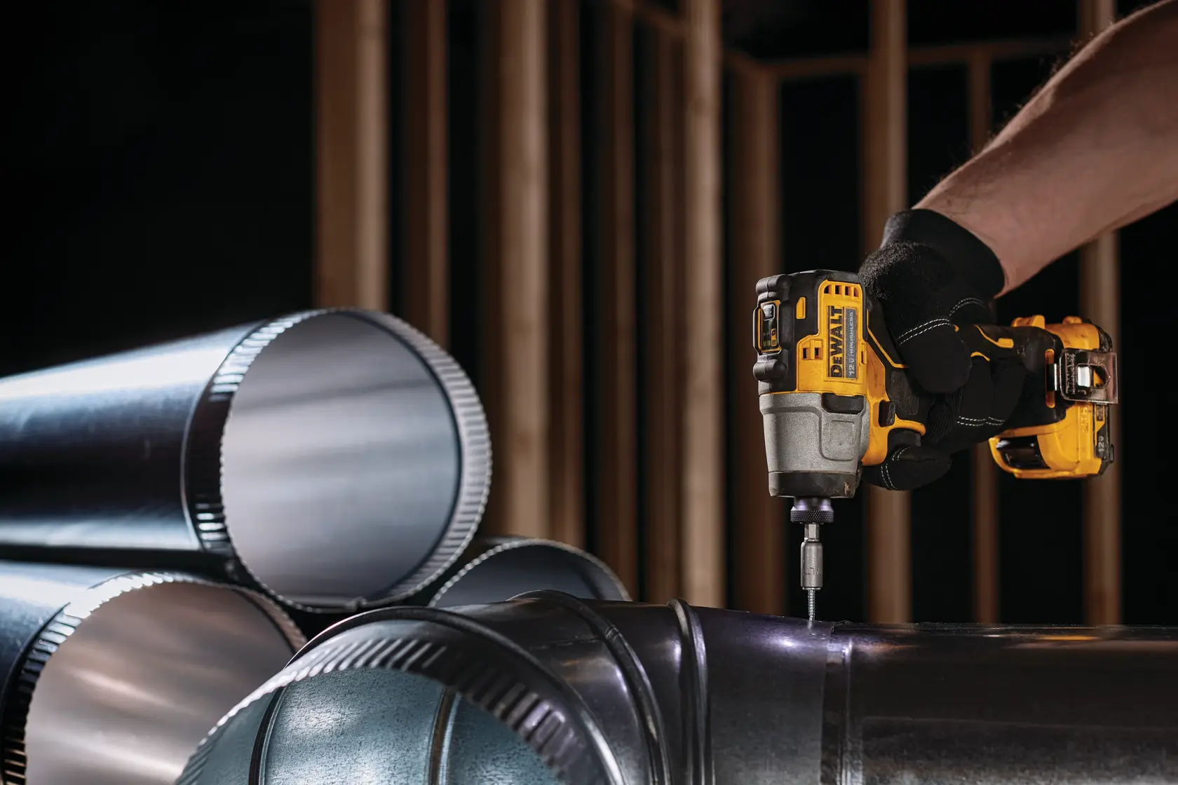 A person wearing black gloves is using a DEWALT cordless impact driver to fasten a screw into metal ductwork pipes. Several cylindrical metal duct pipes are stacked in the foreground, and wooden framing is visible in the background.