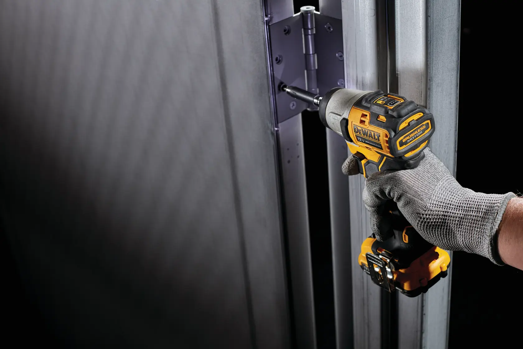 A hand wearing a gray glove uses a DEWALT cordless impact driver to tighten screws on a metal hinge attached to a metal frame.