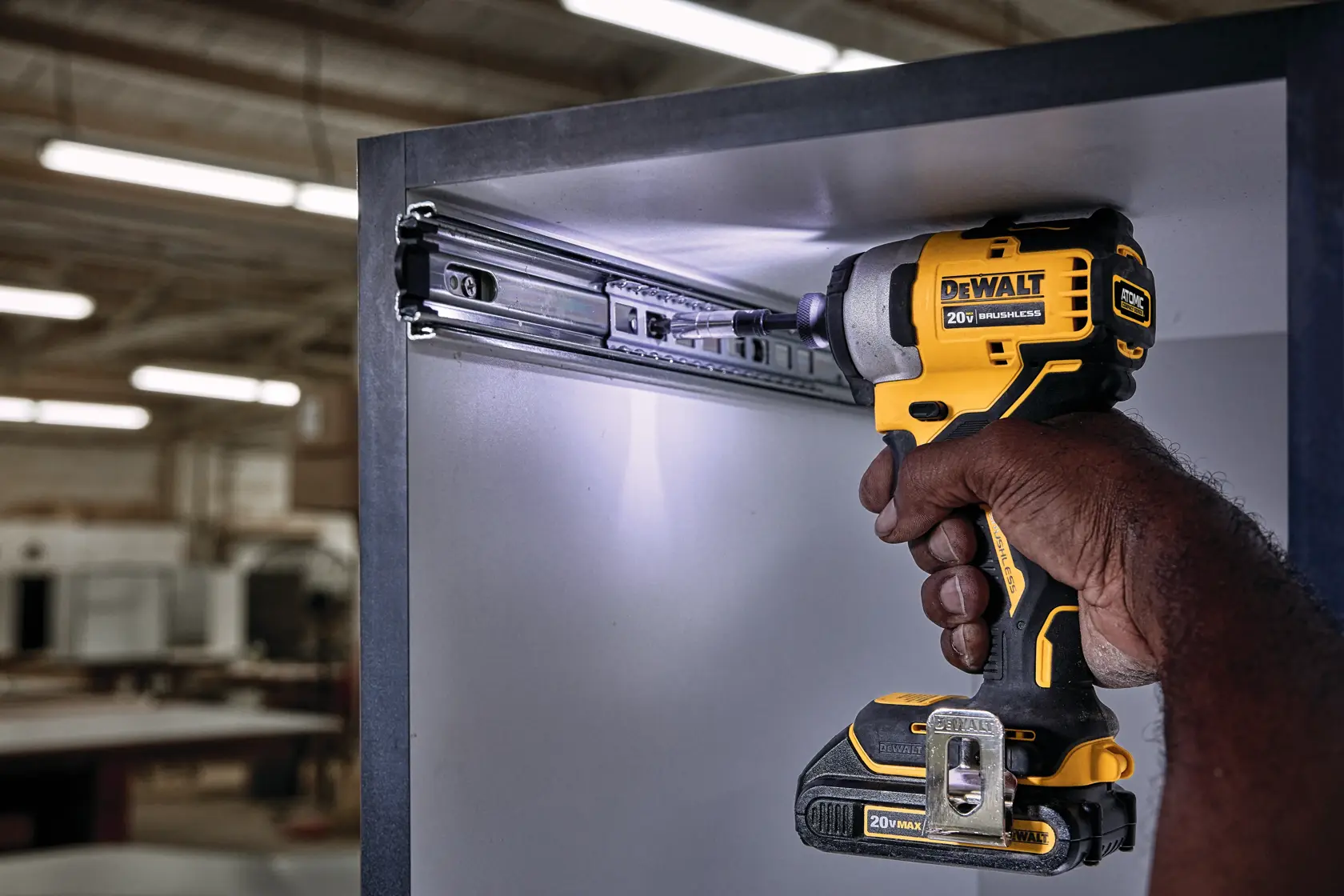 A person using a DEWALT 20V cordless impact driver to install or adjust a metal drawer slide inside a cabinet, in a workshop setting.