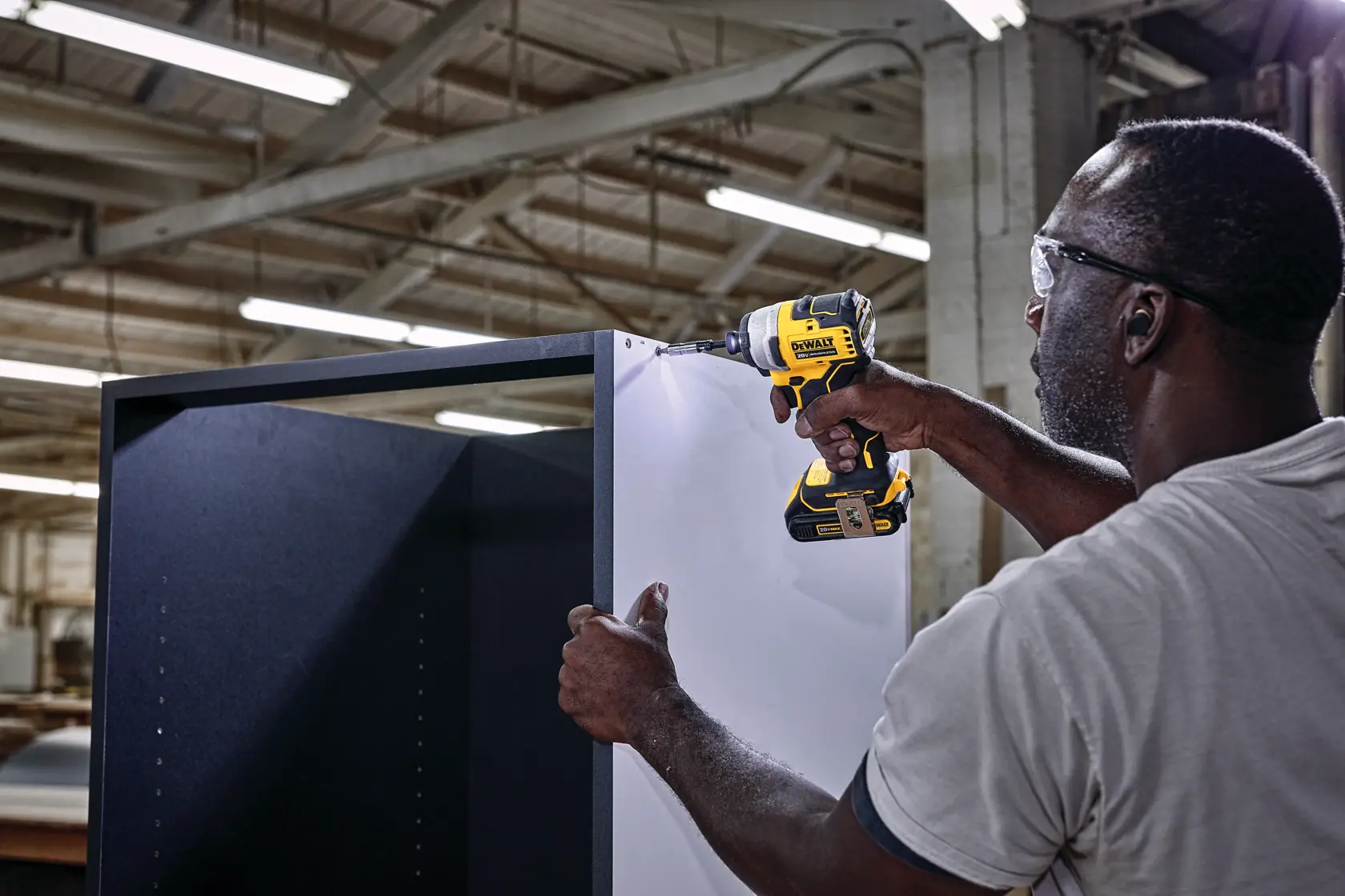 A person uses a DEWALT cordless power drill, model DCF809B, to assemble a large cabinet inside an industrial workshop with visible lighting and beams overhead.