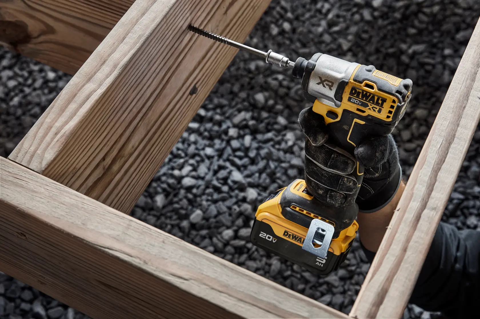A person wearing gloves is using a DEWALT 20V XR power drill with a drill bit to make a hole in a wooden frame at a construction site with gravel on the ground.