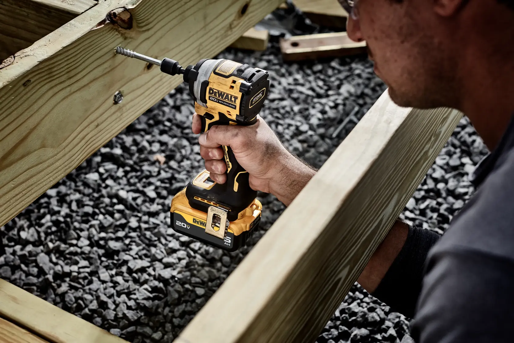 A person uses a DEWALT cordless impact driver to drive a screw into a wooden beam during outdoor construction. The tool is identified as a DEWALT 20V model DCF850P1.