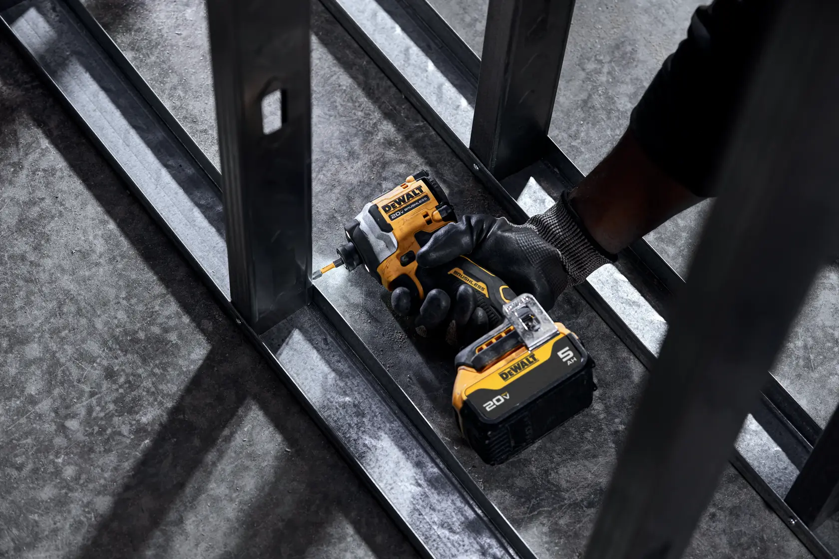 A person wearing a black glove uses a DEWALT 20V cordless impact driver to drive a screw into a metal frame on a concrete floor.