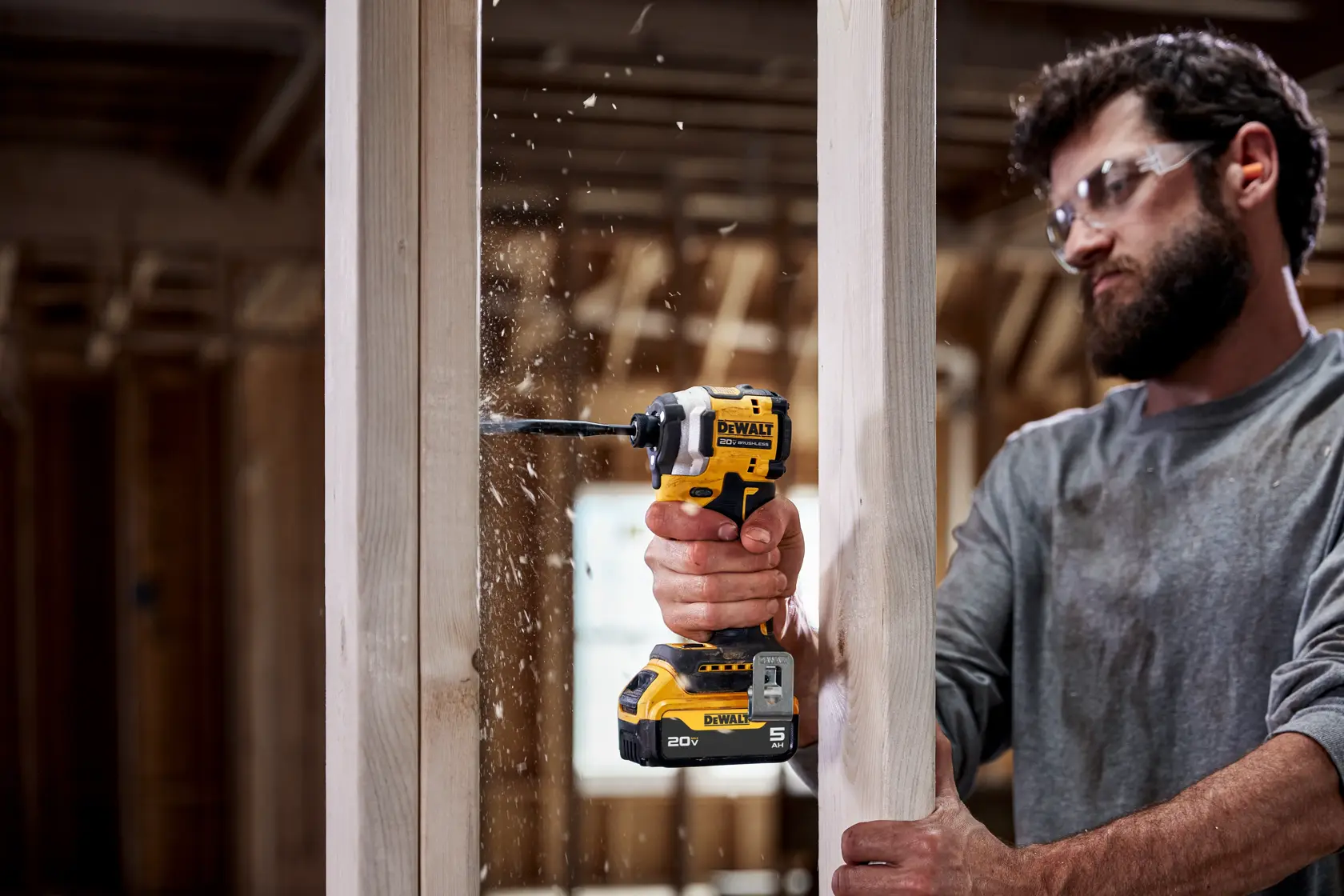 A person using a DEWALT 20V cordless drill (model DCF850P1) to drill into a wooden frame in a construction setting, with wood chips flying out. The person's face is blurred for privacy.
