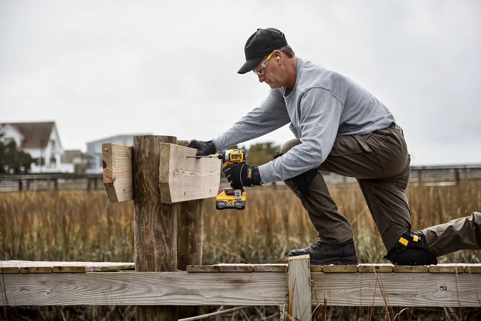 A worker wearing gloves uses a DEWALT cordless drill (model DCF860WW1) to fasten wooden boards to a post outdoors. He is kneeling on a wooden structure, with a grassy area and houses visible in the background.