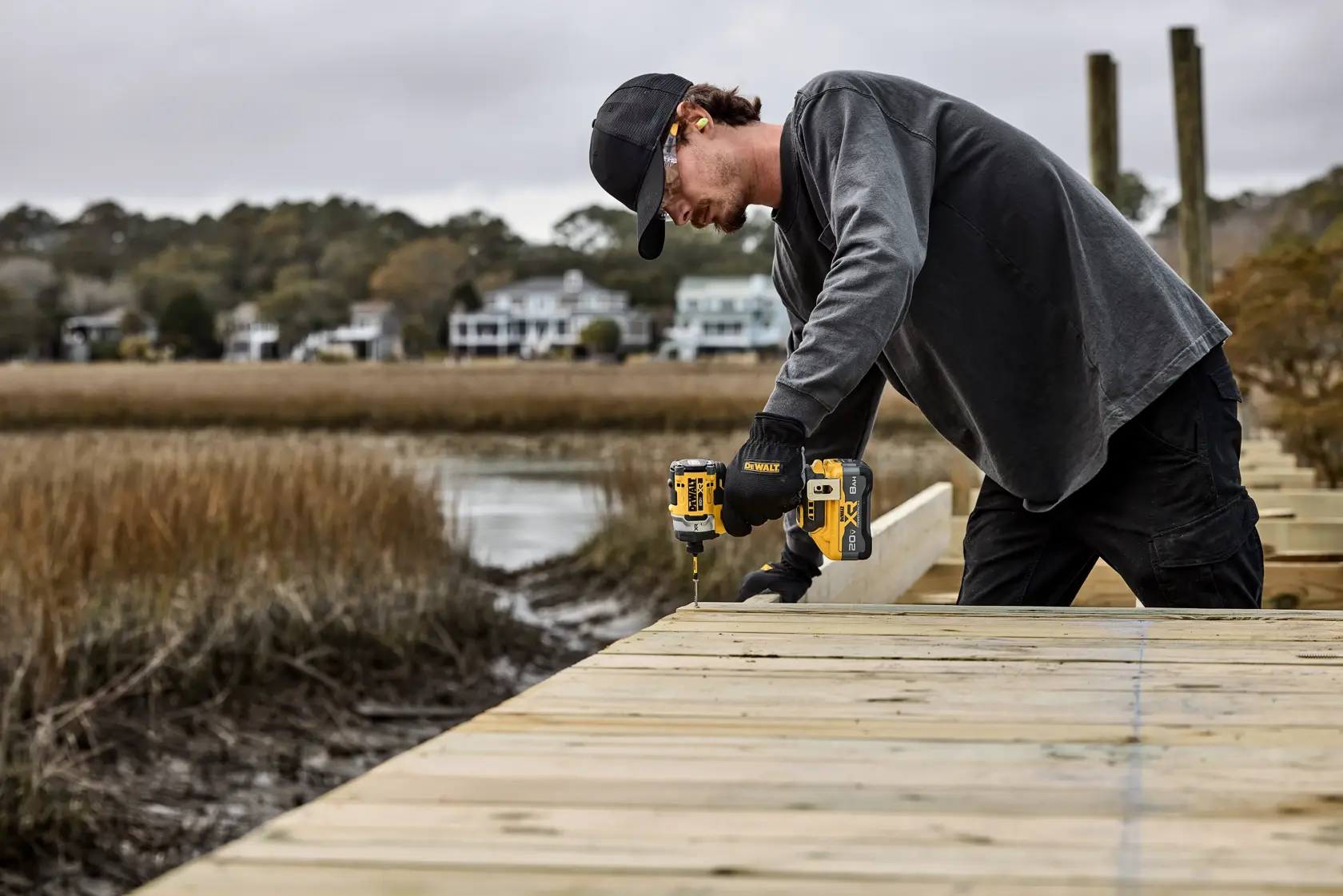 A person using a DEWALT cordless power drill to work on an outdoor wooden deck construction project near a body of water. The person is wearing a black cap, gloves, and gray long-sleeve shirt.