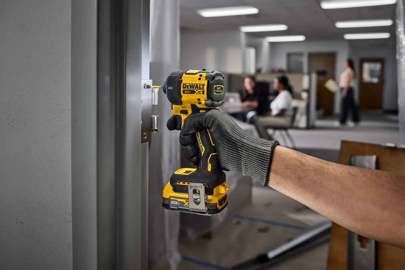 A person wearing work gloves is using a DEWALT cordless impact wrench (model DCF870B) to install or tighten a bolt on a metal door frame in an indoor workspace.