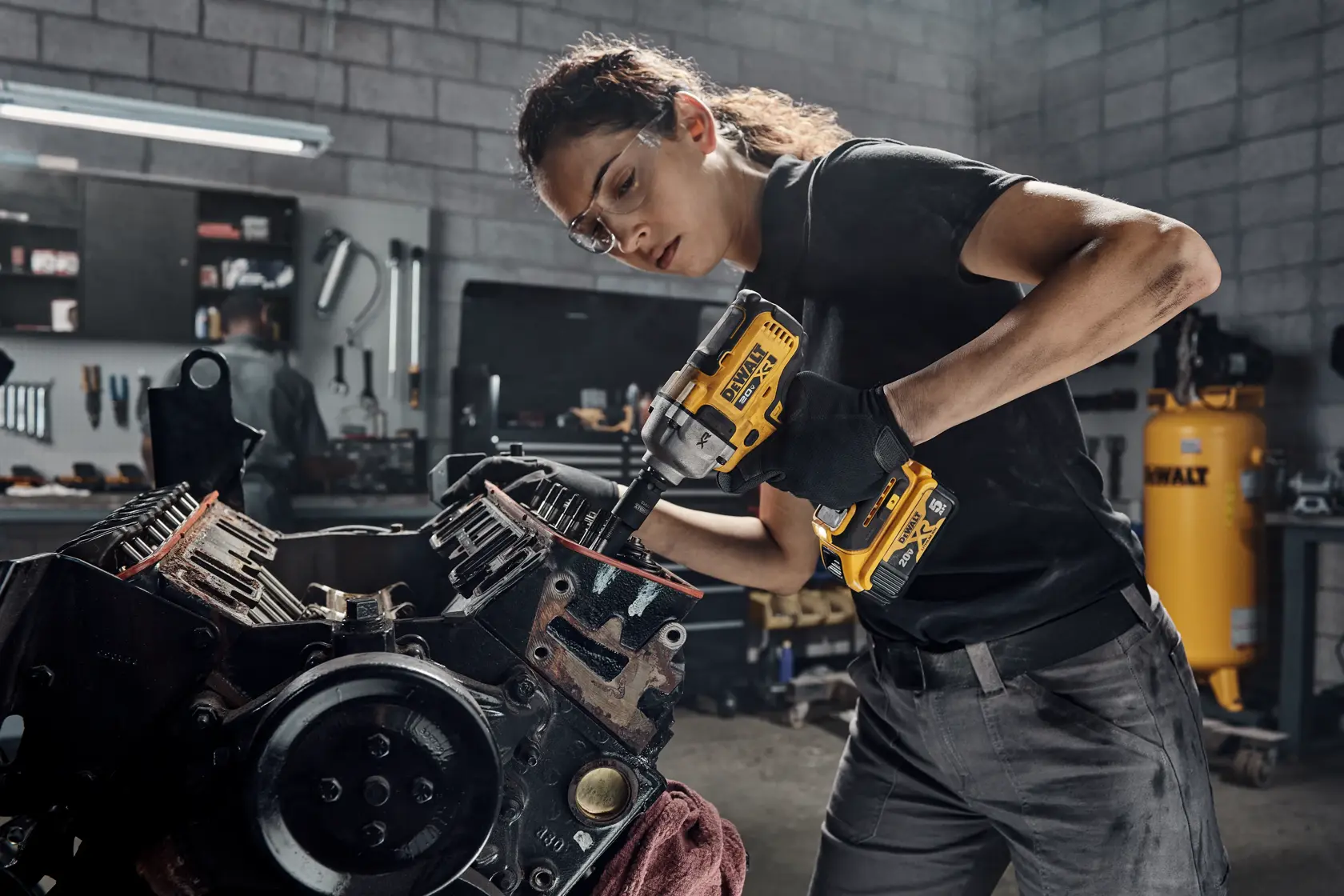 Person using a DEWALT DCF891B cordless impact wrench to work on an engine in a workshop. Their face is blurred for privacy. The workshop background includes tools and a DEWALT air compressor.